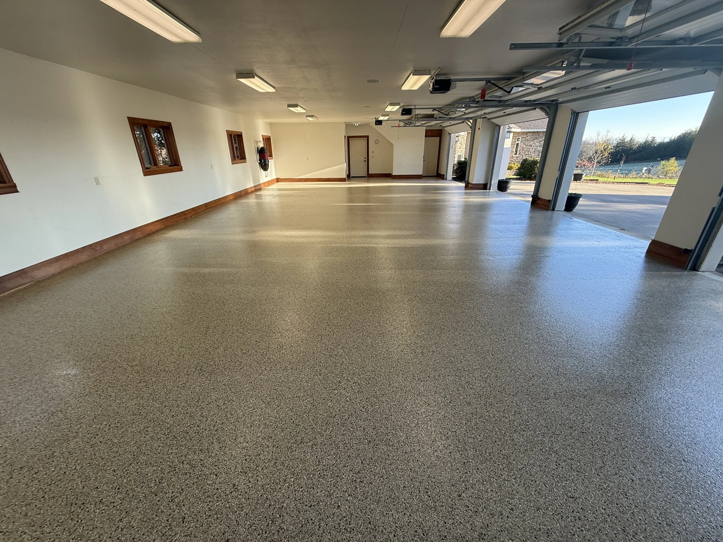 Empty garage with a polished speckled concrete floor, white walls, wooden trim, small windows, and an open roll-up door leading outside.