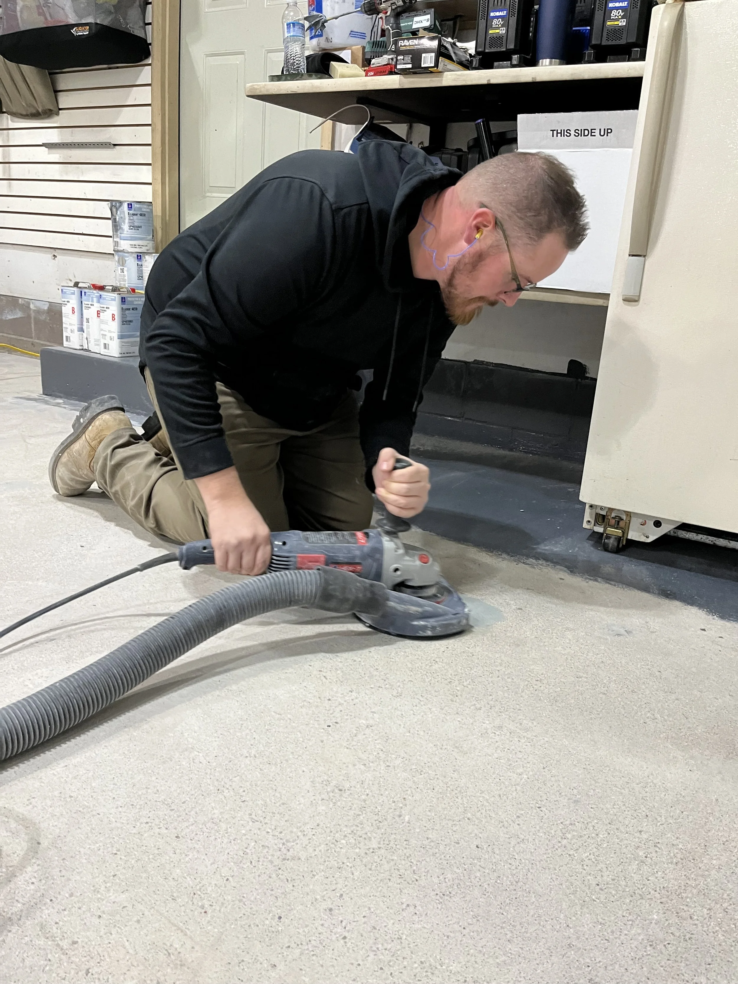 Man kneeling on the floor using a sander on a concrete in a workshop or garage.
