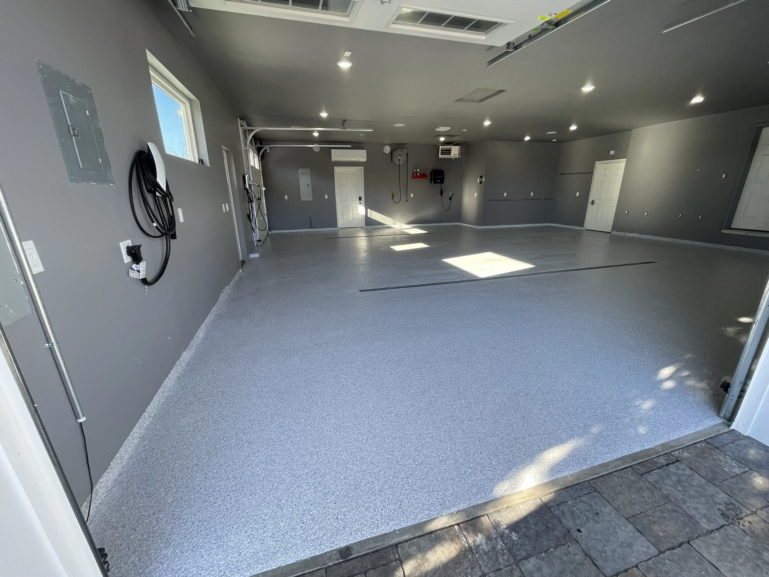 Empty interior garage with gray walls, polished concrete floors, and multiple windows. There are electrical outlets, a wall-mounted telephone, and utility panels. Garage doors are open, revealing a paved driveway outside. The ceiling has recessed lig