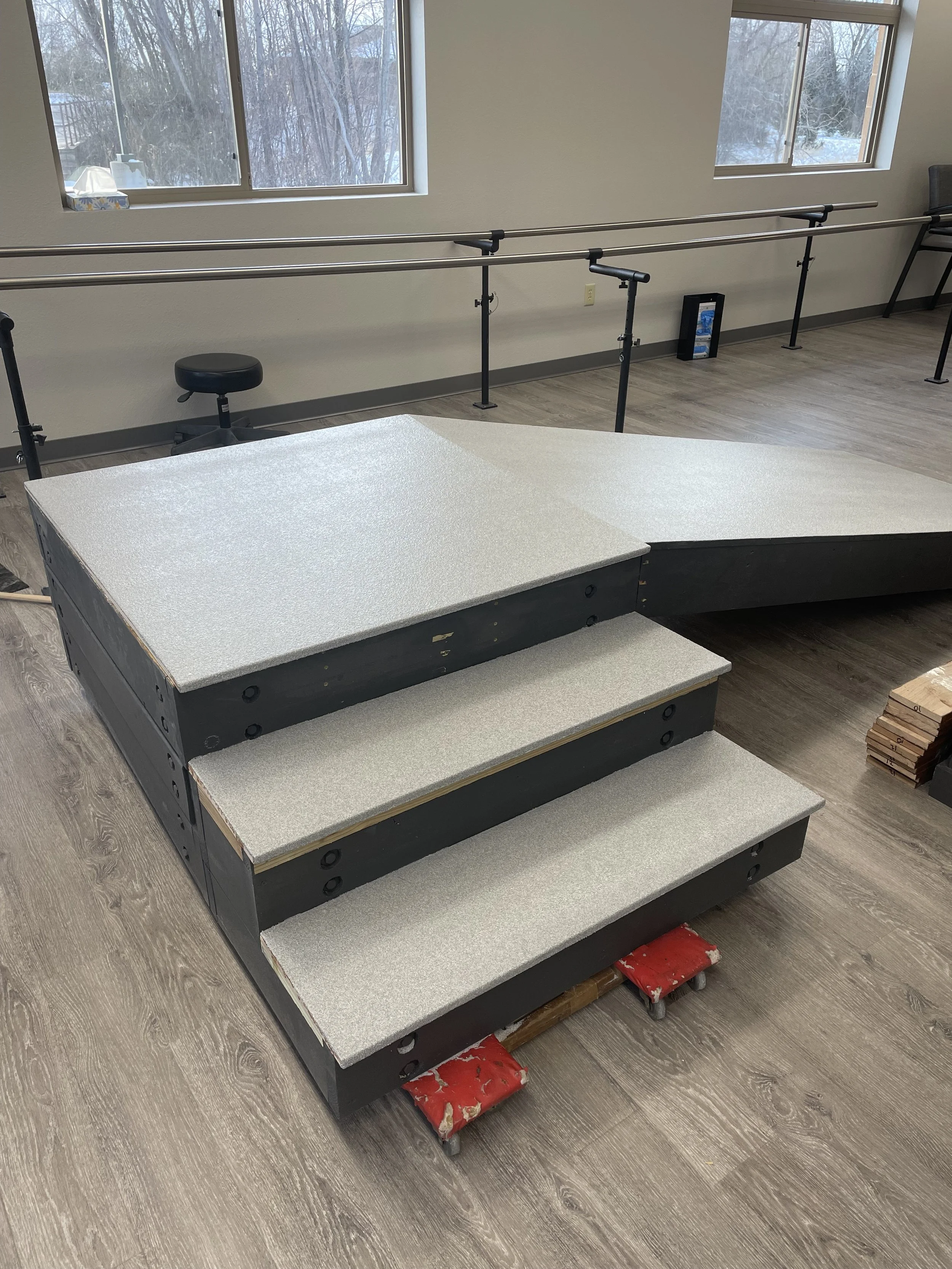 Dance studio with a portable stage platform, ballet barres along the wall, wooden flooring, and a window showing a snowy outdoor scene.