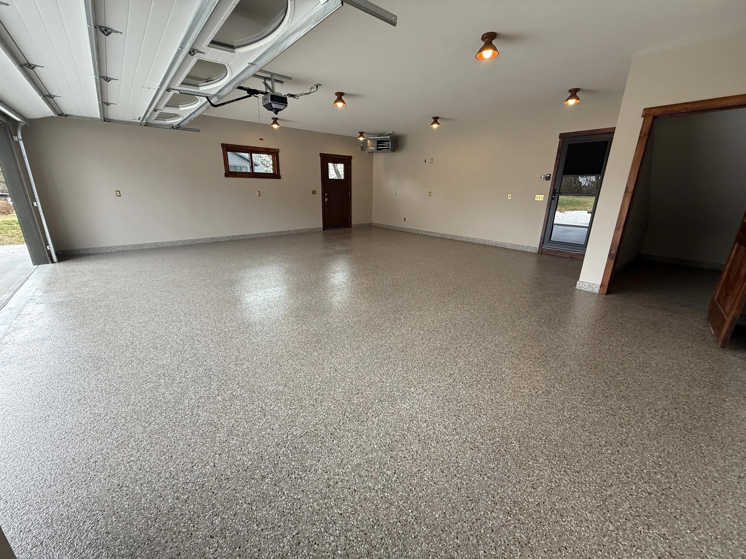An empty spacious garage with speckled epoxy flooring, white walls, small window, garage door, and a side door leading outside.