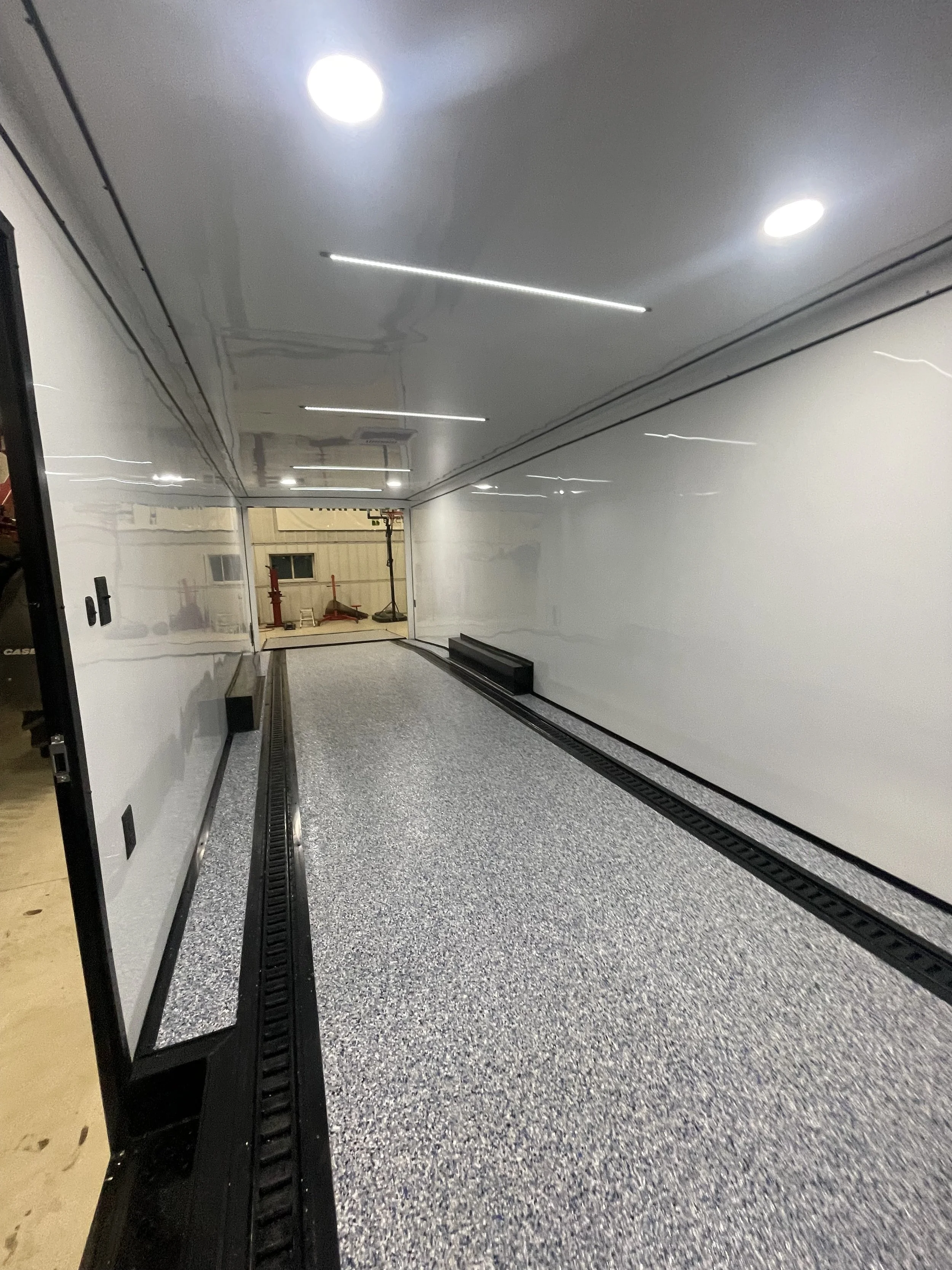 Empty enclosed trailer with a speckled gray floor, white walls, and bright ceiling lights.