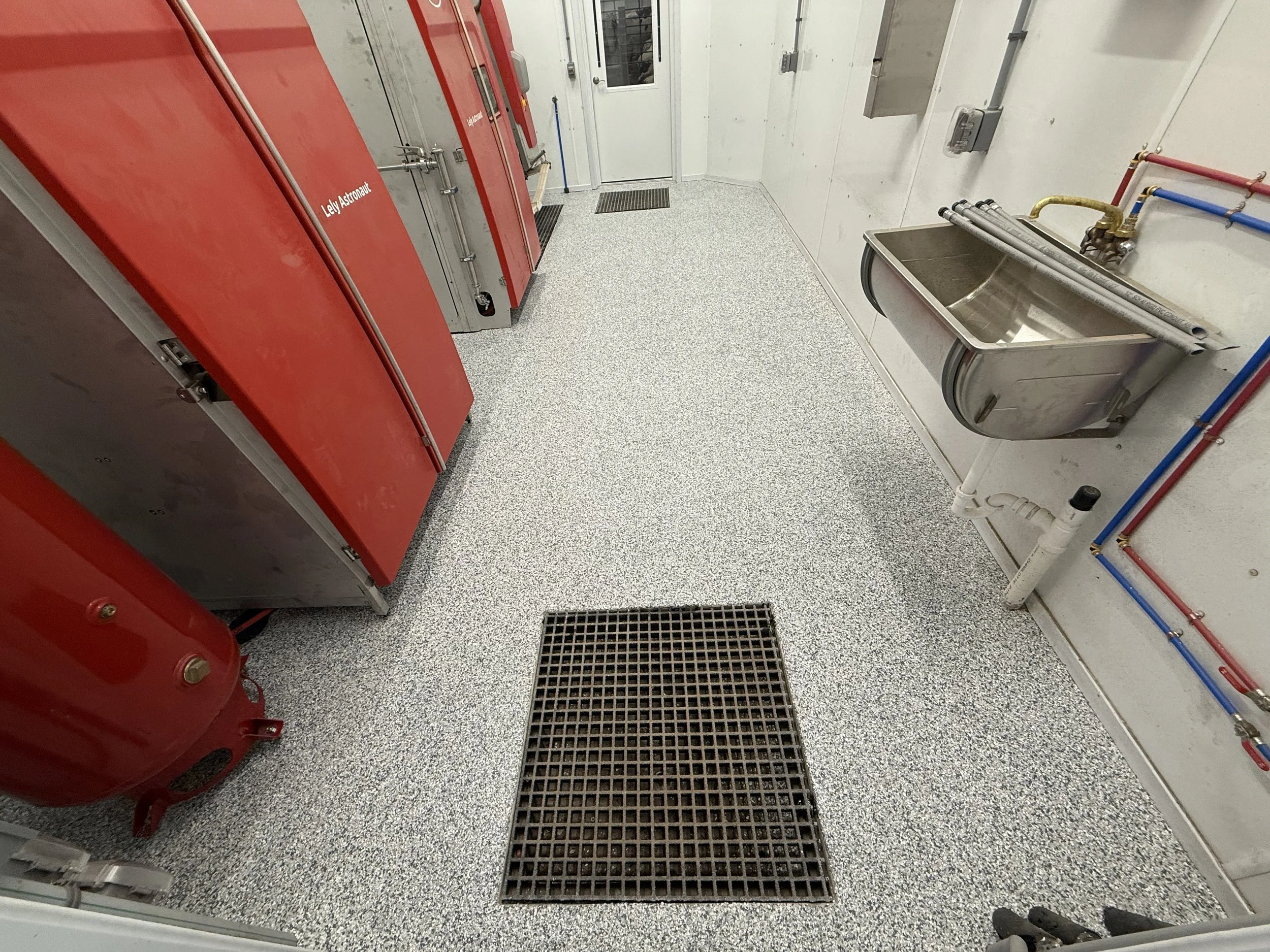 A utility room with red fire safety equipment, a stainless steel sink with pipes, a floor drain, and various colored pipes on the wall.