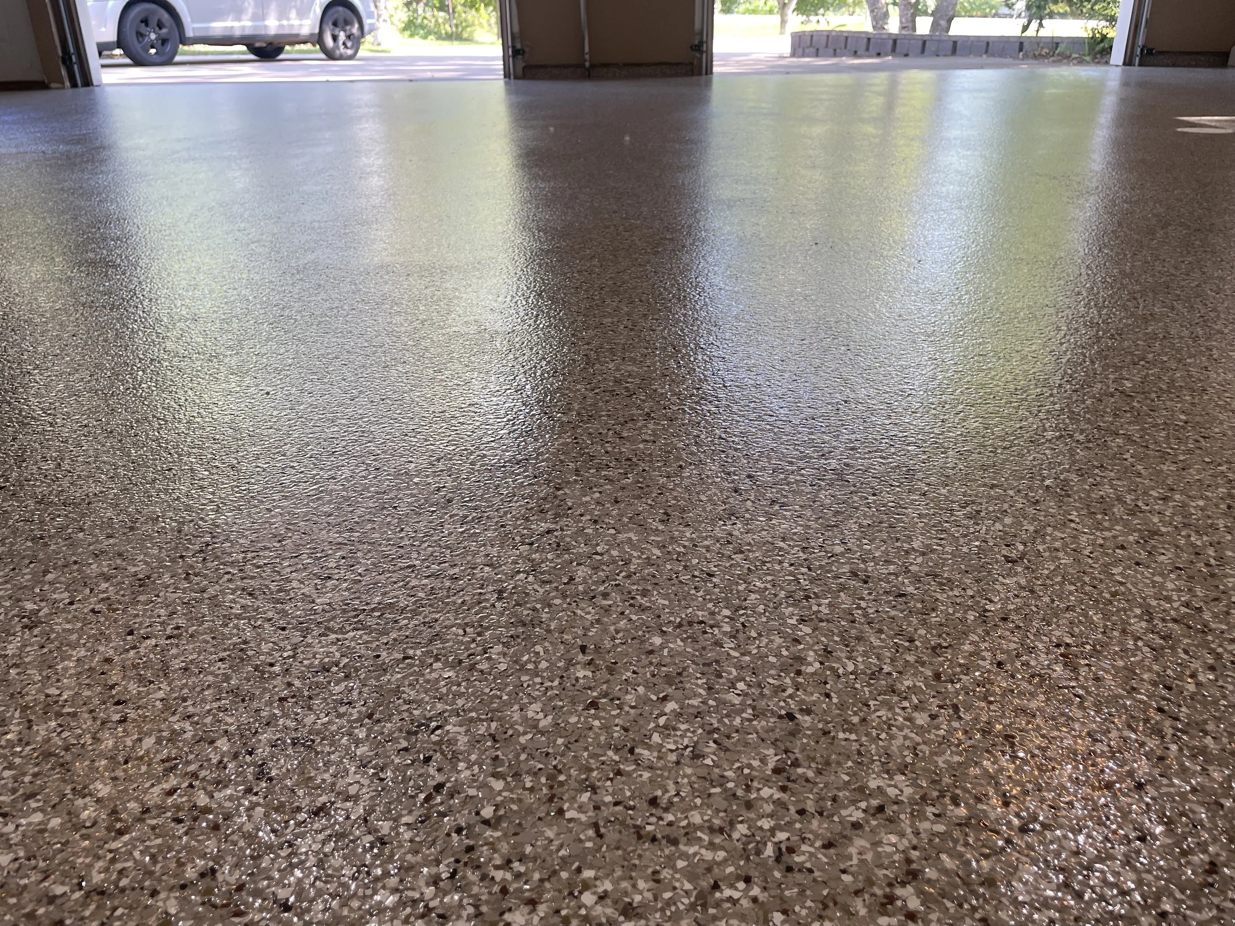 Smooth, polished, speckled epoxy garage floor with a view of the open garage door, driveway, parked vehicle, and greenery outside.