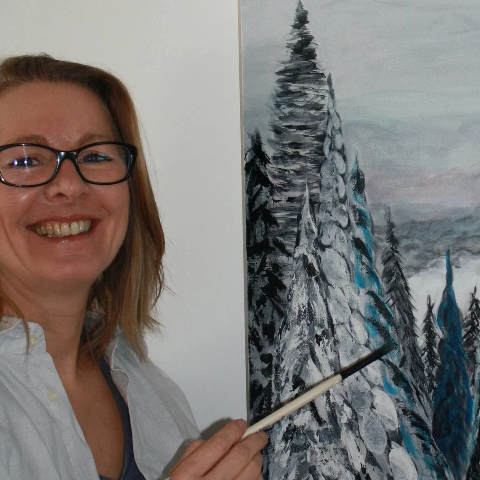 A woman with glasses smiling while holding a paintbrush, standing next to a painting of snow-covered trees on a mountain. The painting features black, white, and blue colors.