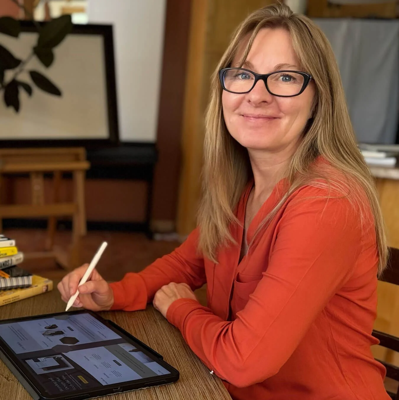 A woman with long blonde hair, wearing glasses and an orange blouse, sitting at a wooden table using a stylus on a tablet.