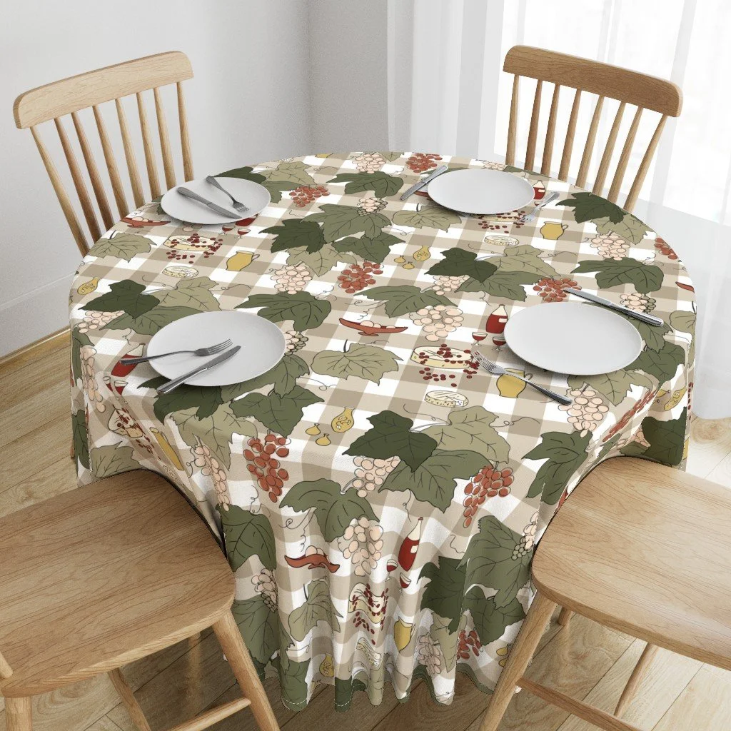 A round dining table covered with a holiday-themed tablecloth with leaves, grapes, and wine bottles, set with four white plates and silverware, surrounded by four wooden chairs in a bright room.