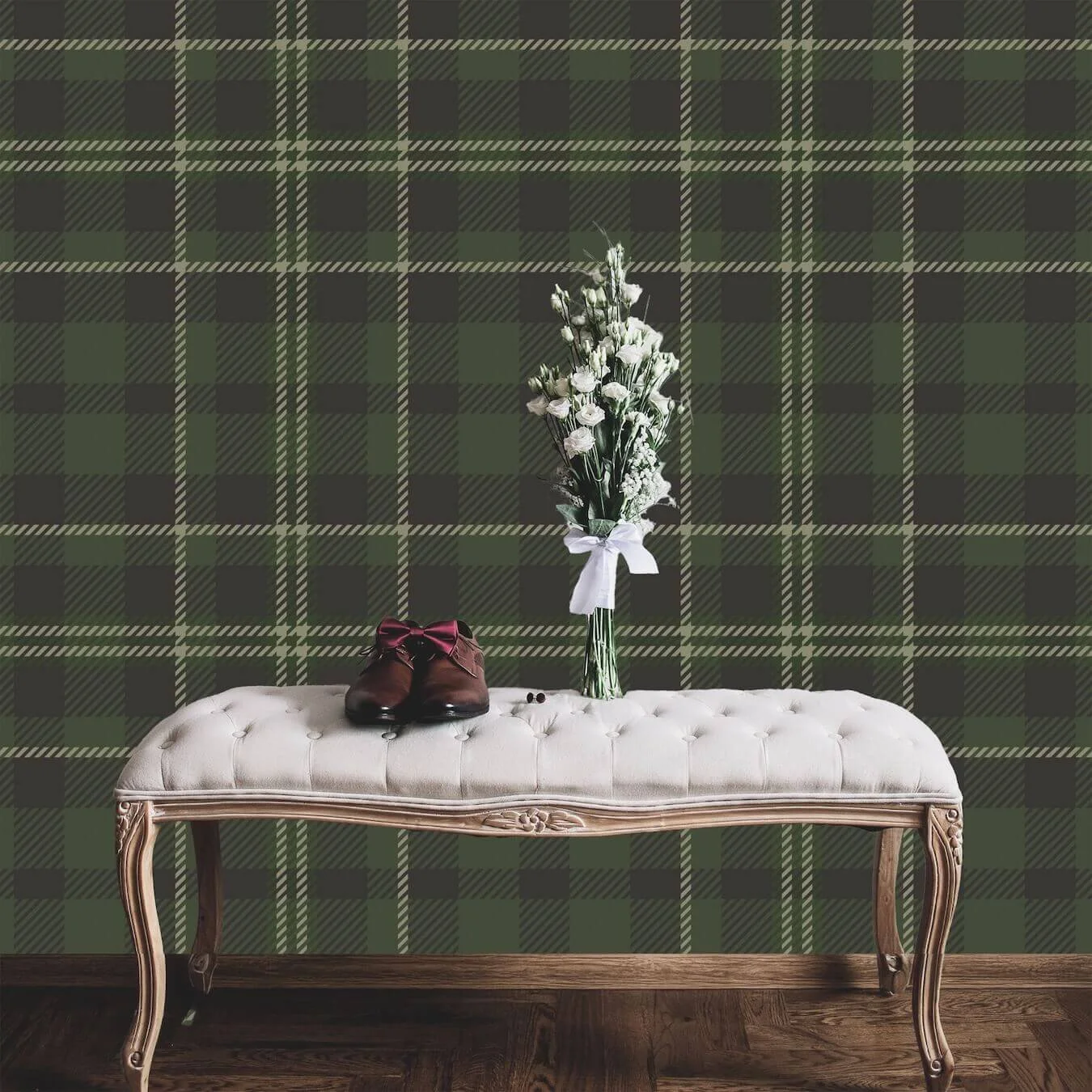 A beige upholstered bench with wooden carved legs, on a hardwood floor, against a green and dark gray plaid wallpaper. On the bench, there are brown shoes with pink laces, a glass vase with white flowers tied with a white ribbon, and two small dark o