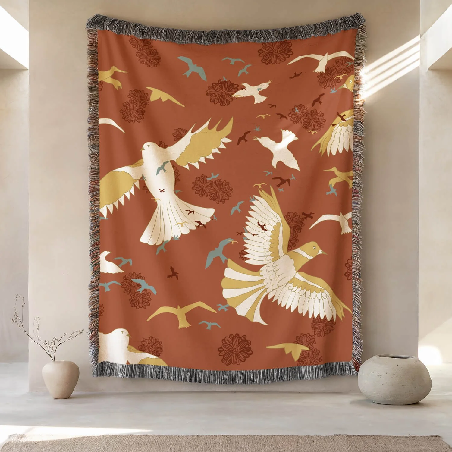 A large textile wall hanging with a warm terracotta background depicting white and gold birds flying among red floral patterns, bordered with gray fringe, hanging on a wall between two columns with sunlight streaming through.