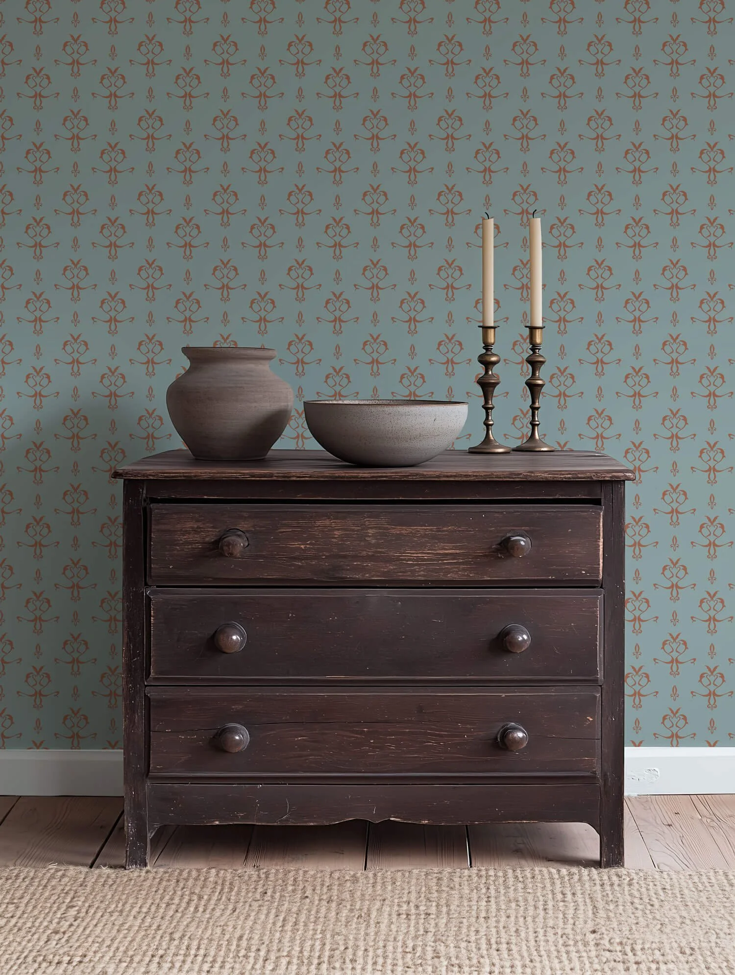 A dark wooden dresser with four drawers and wooden knobs. On top of the dresser are two candle holders with white candles, a large ceramic vase, and a shallow ceramic bowl. The dresser is against a wall with light blue wallpaper that has an orange fl