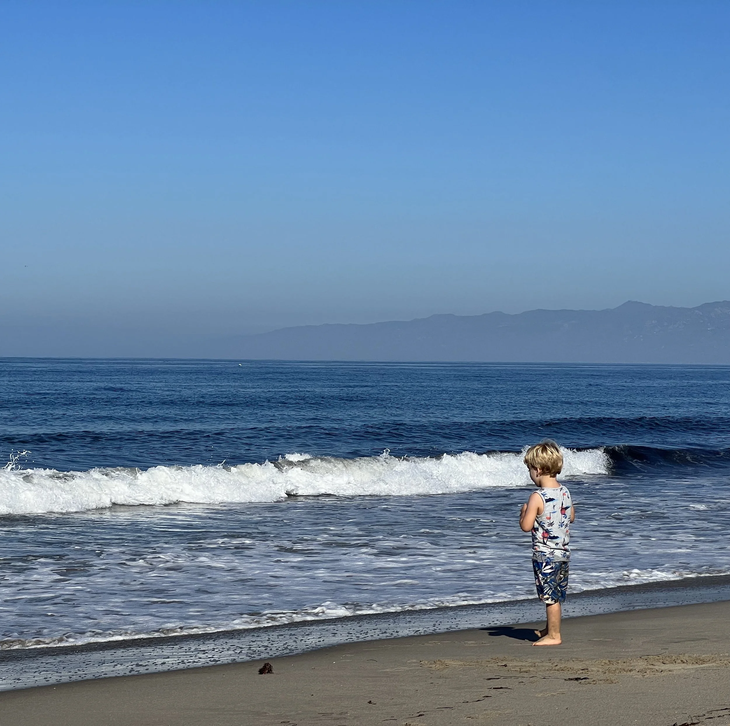 A young boy with blond hair wearing a tank top and shorts stands on the sandy beach near the shoreline, watching the ocean waves under a clear blue sky with distant mountains in the background.
