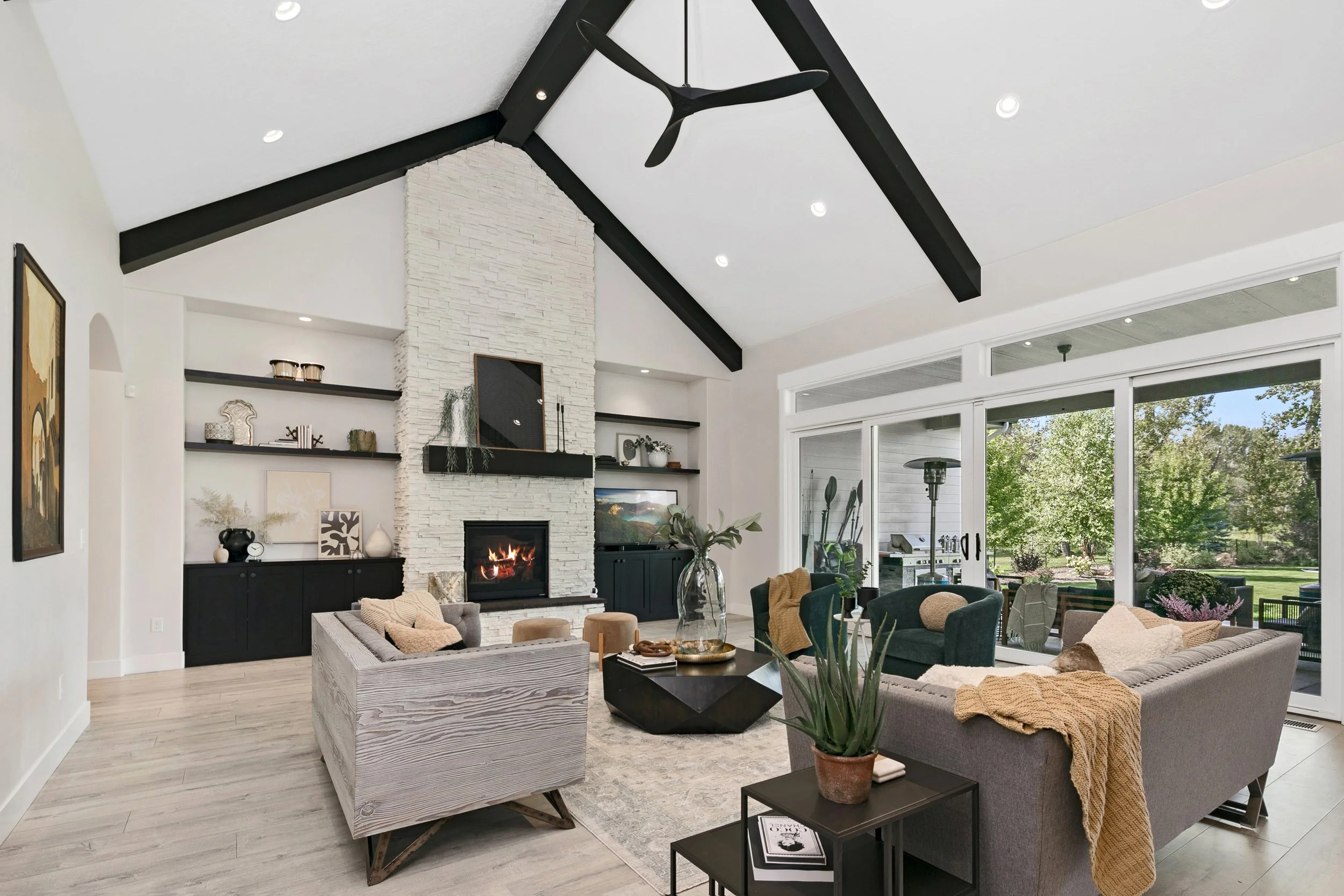 Living room with a white fireplace, black built-in cabinets, and open shelves, a ceiling fan, large sliding glass doors leading to an outdoor patio, and modern furniture including a gray sofa, an armchair, and a black coffee table.