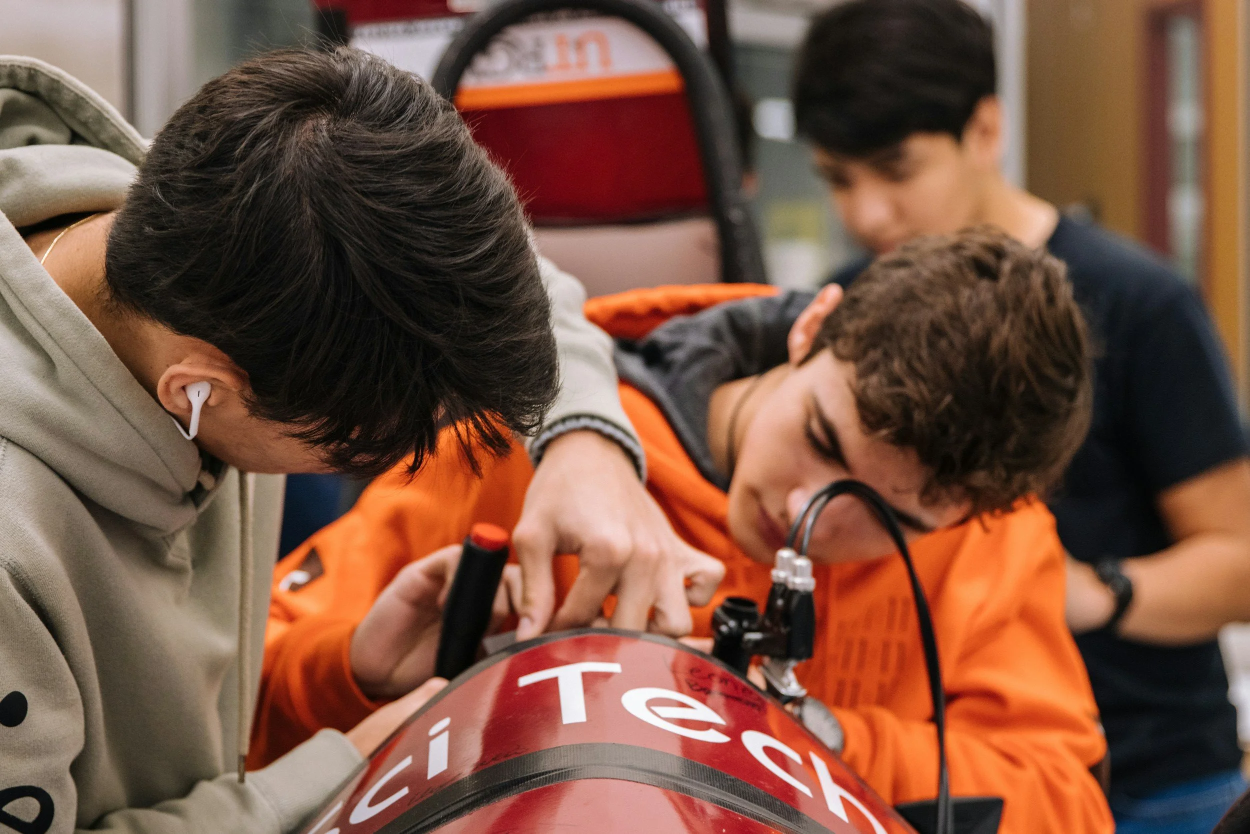 Three young men working on a red and white project, with one using a soldering iron and the other two observing closely.