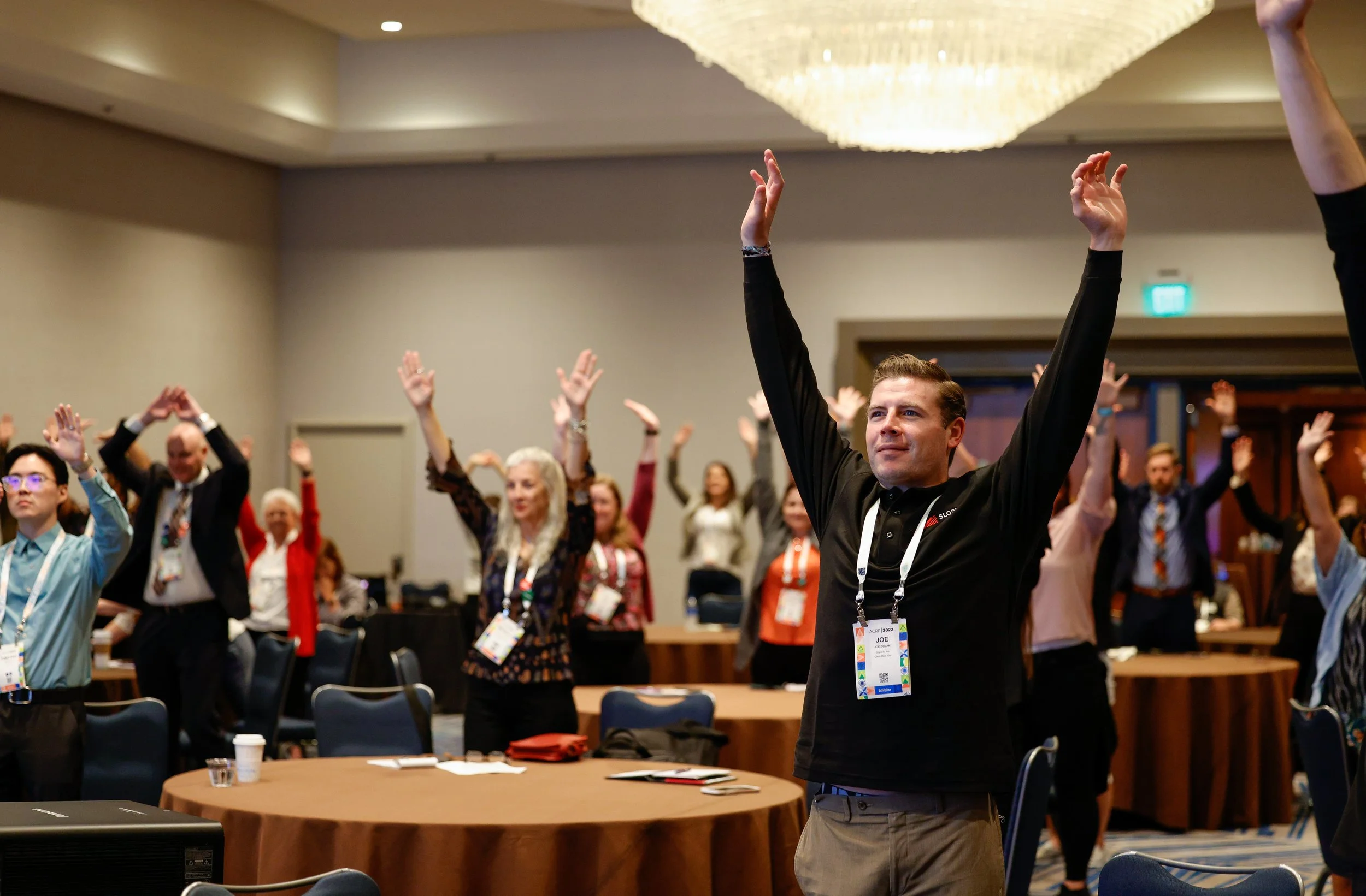 People participating in a group activity at a conference, standing with arms raised in a room with round tables and a large chandelier.