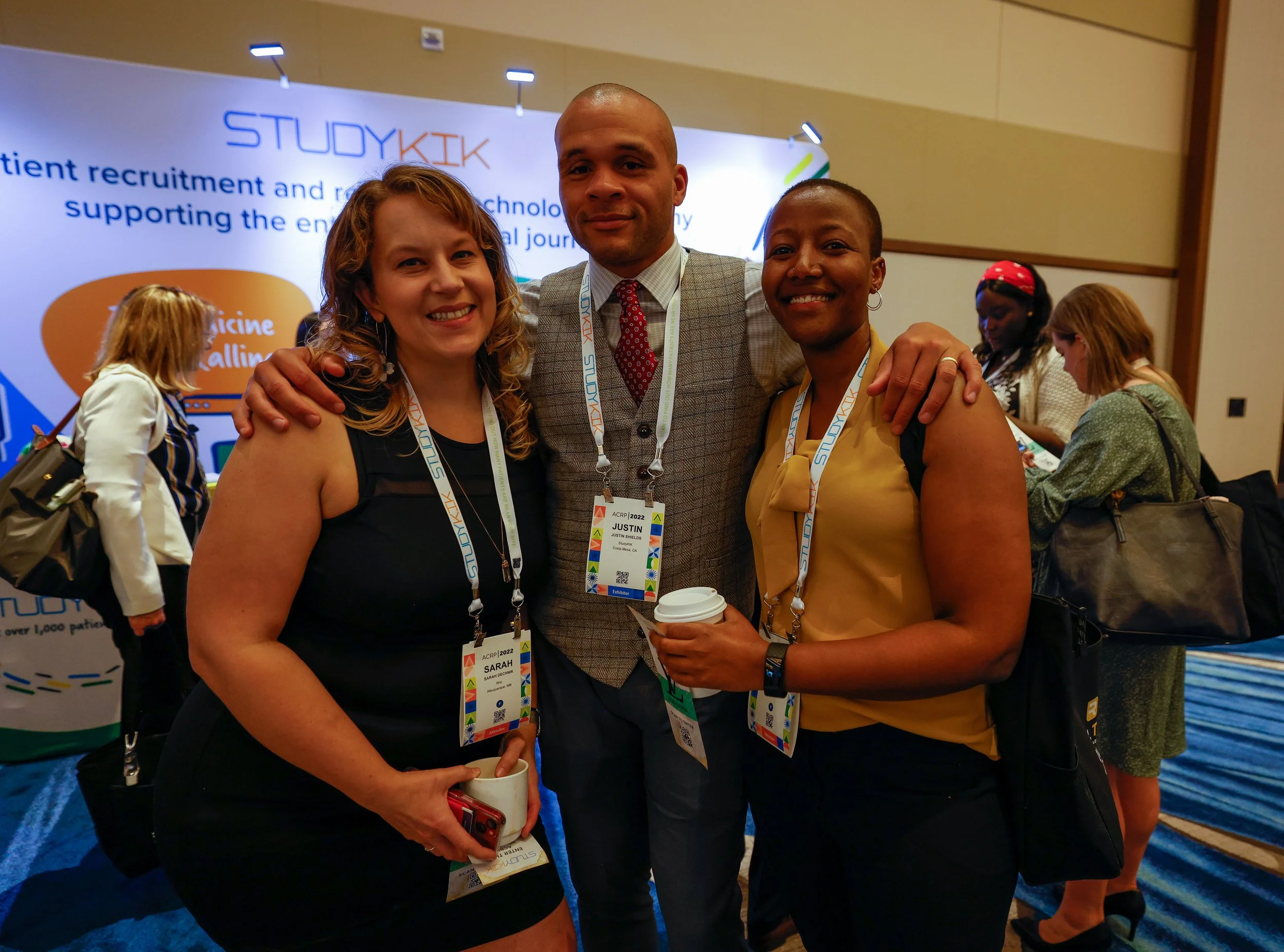 Three smiling conference attendees with name badges, standing close together with arms around each other, at a professional event.