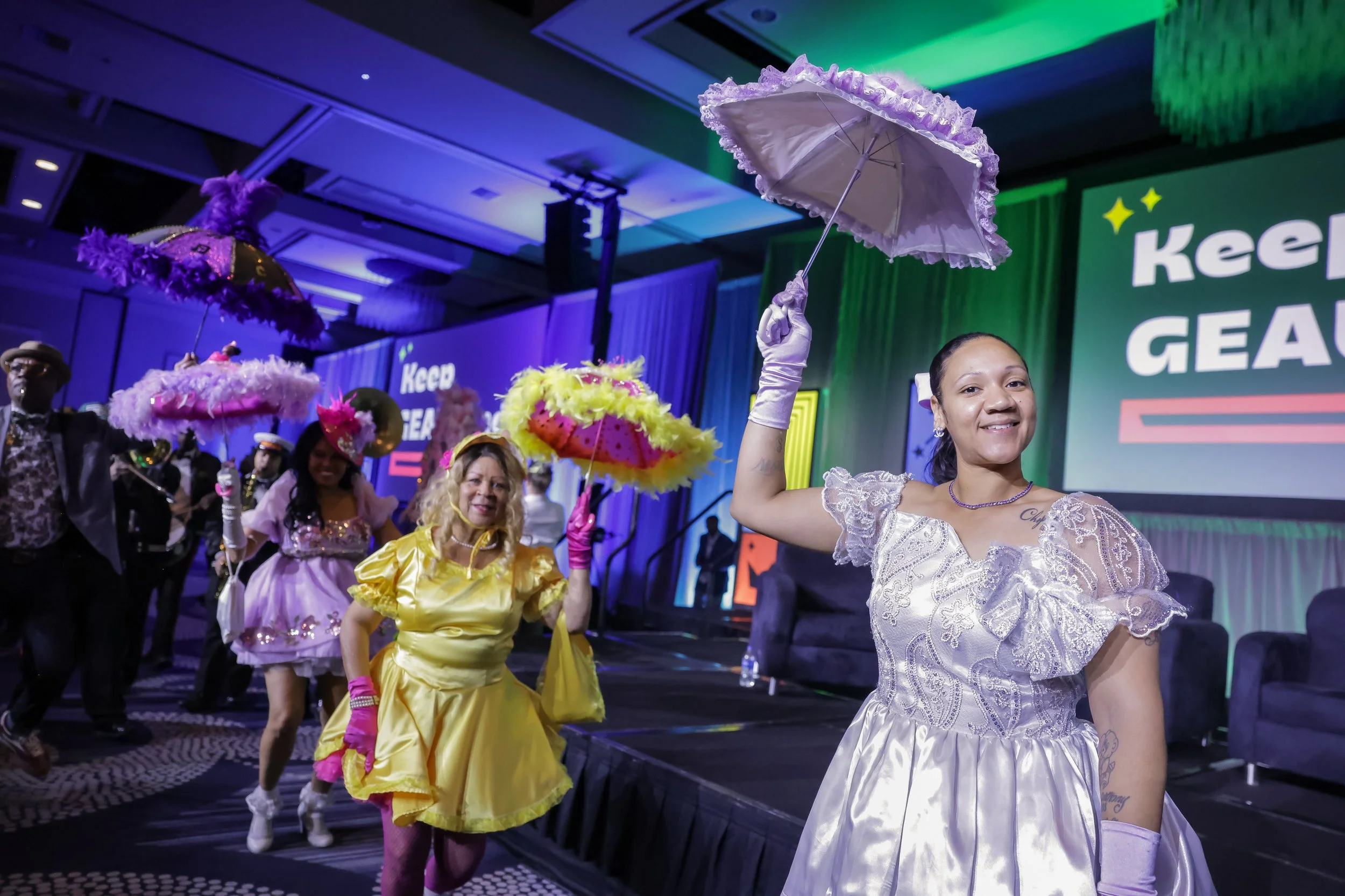 People dressed in colorful vintage costumes with parasols participating in a lively event or parade indoors with stage and large screens in background.