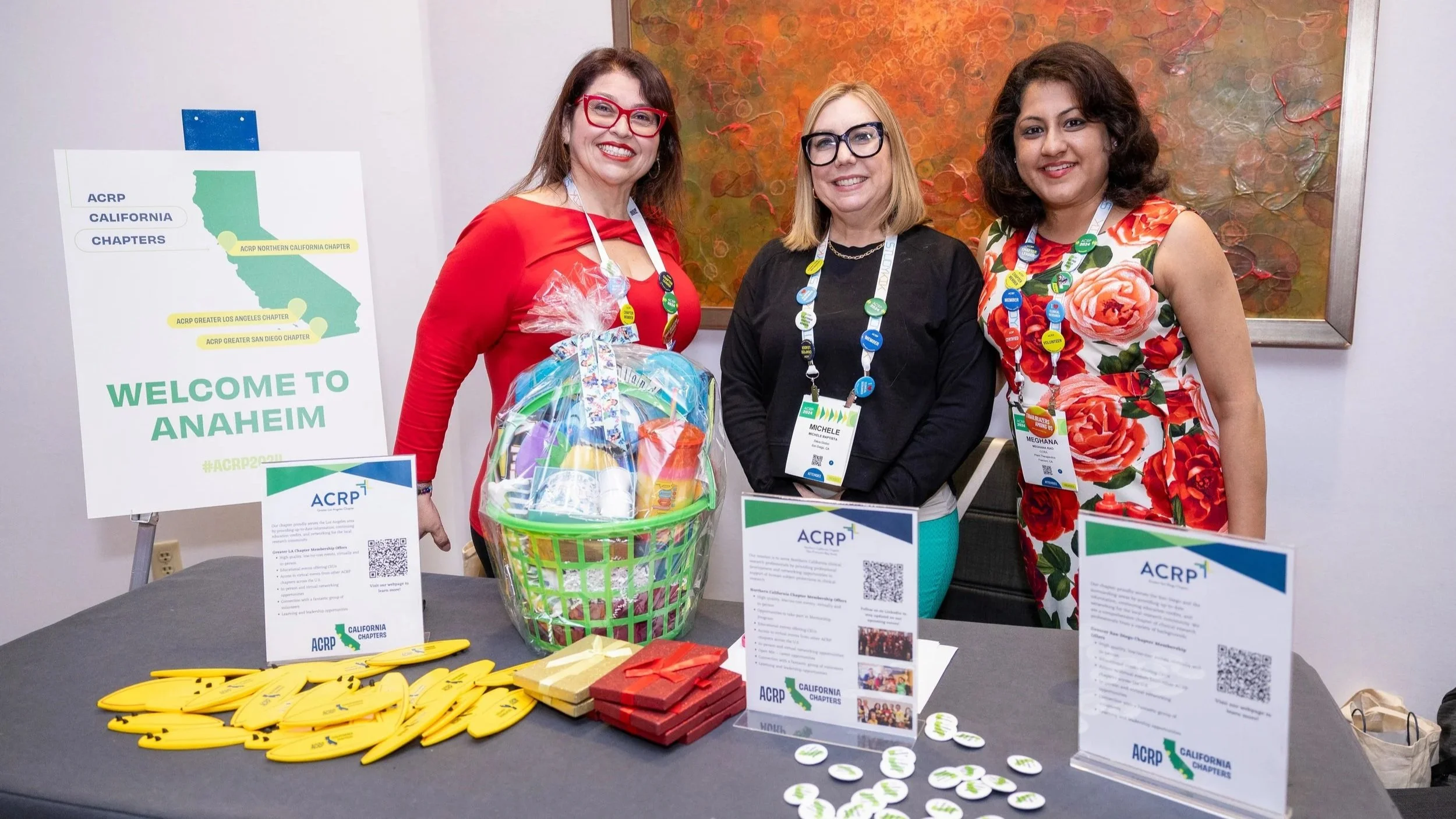 Three women standing behind a table at a conference, smiling and wearing name badges with buttons. The table has yellow pens, red and gold wrapped gifts, and leaf-shaped items. A basket filled with colorful items is on the table. Behind them, there are signs and an abstract painting.