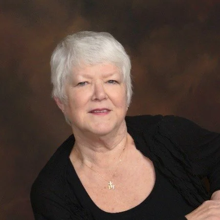 Portrait of an older woman with short white hair wearing a black top and gold necklace, smiling slightly with a dark brown background.