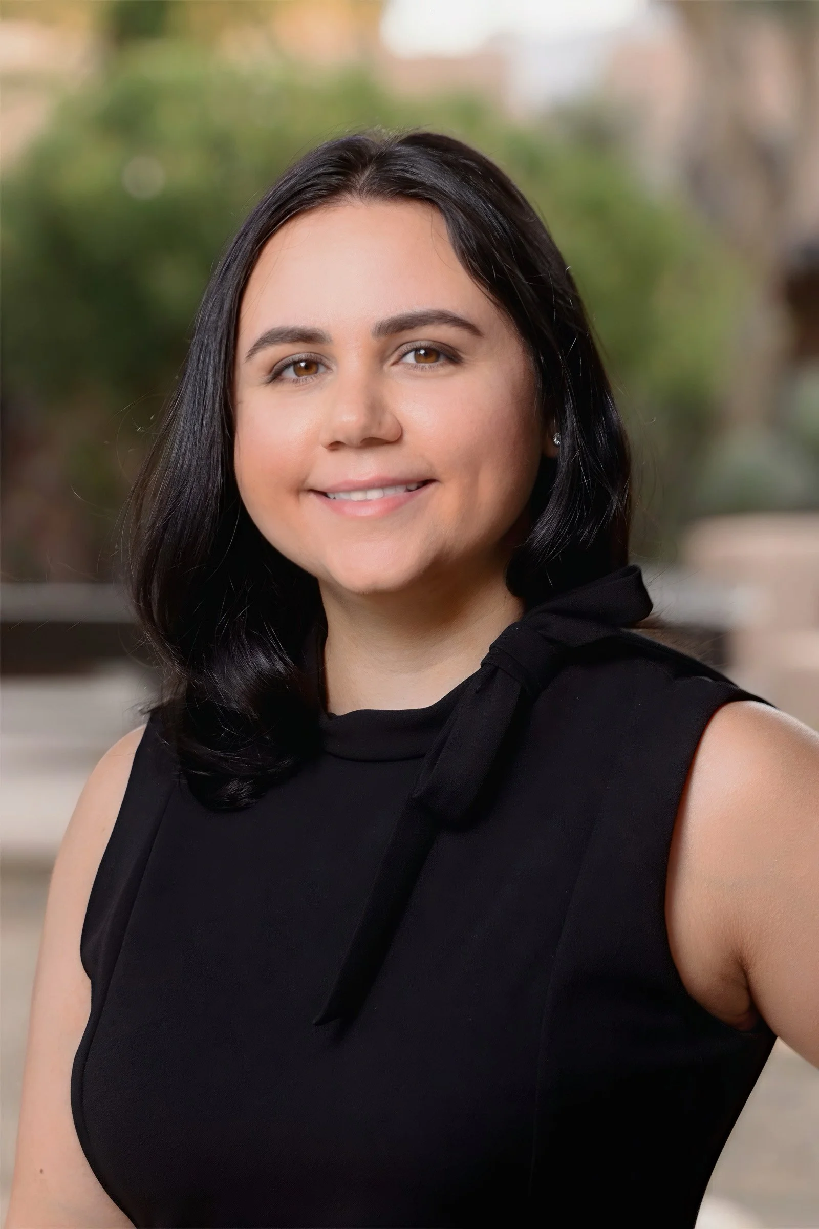 A woman with shoulder-length black hair, wearing a sleeveless black top with a bow detail on the shoulder, smiling outdoors with a blurred green and brown background.