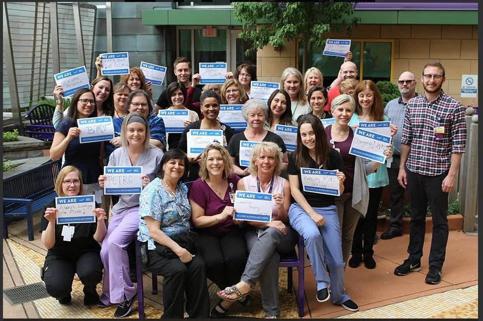 Group of diverse people holding signs reading "We Are" and various associated messages, gathered outdoors in a semi-circle for a photo.