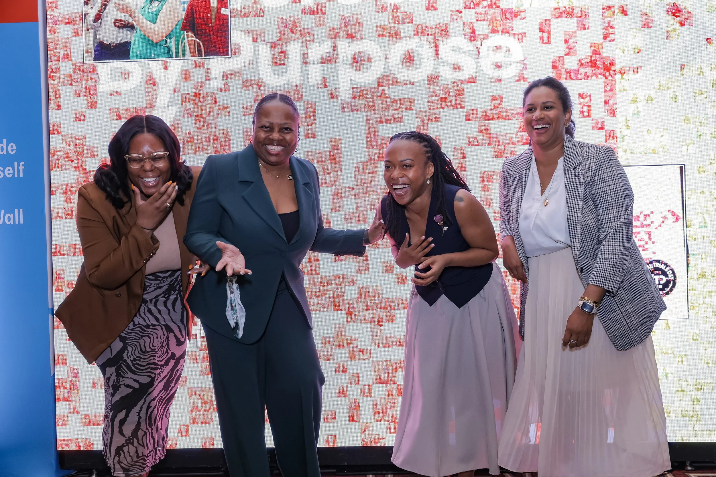 Four women standing together on stage, smiling and laughing, with a digital backdrop and a small photo inset showing two of them.