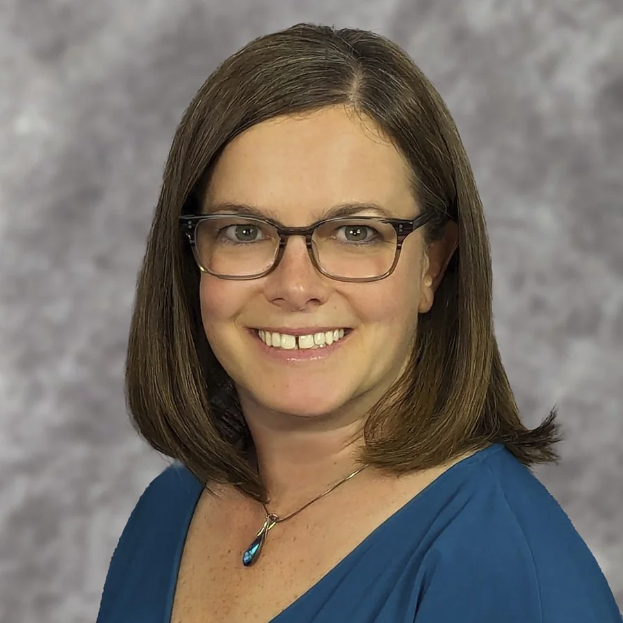 A woman with shoulder-length brown hair and glasses smiling in front of a gray mottled background.