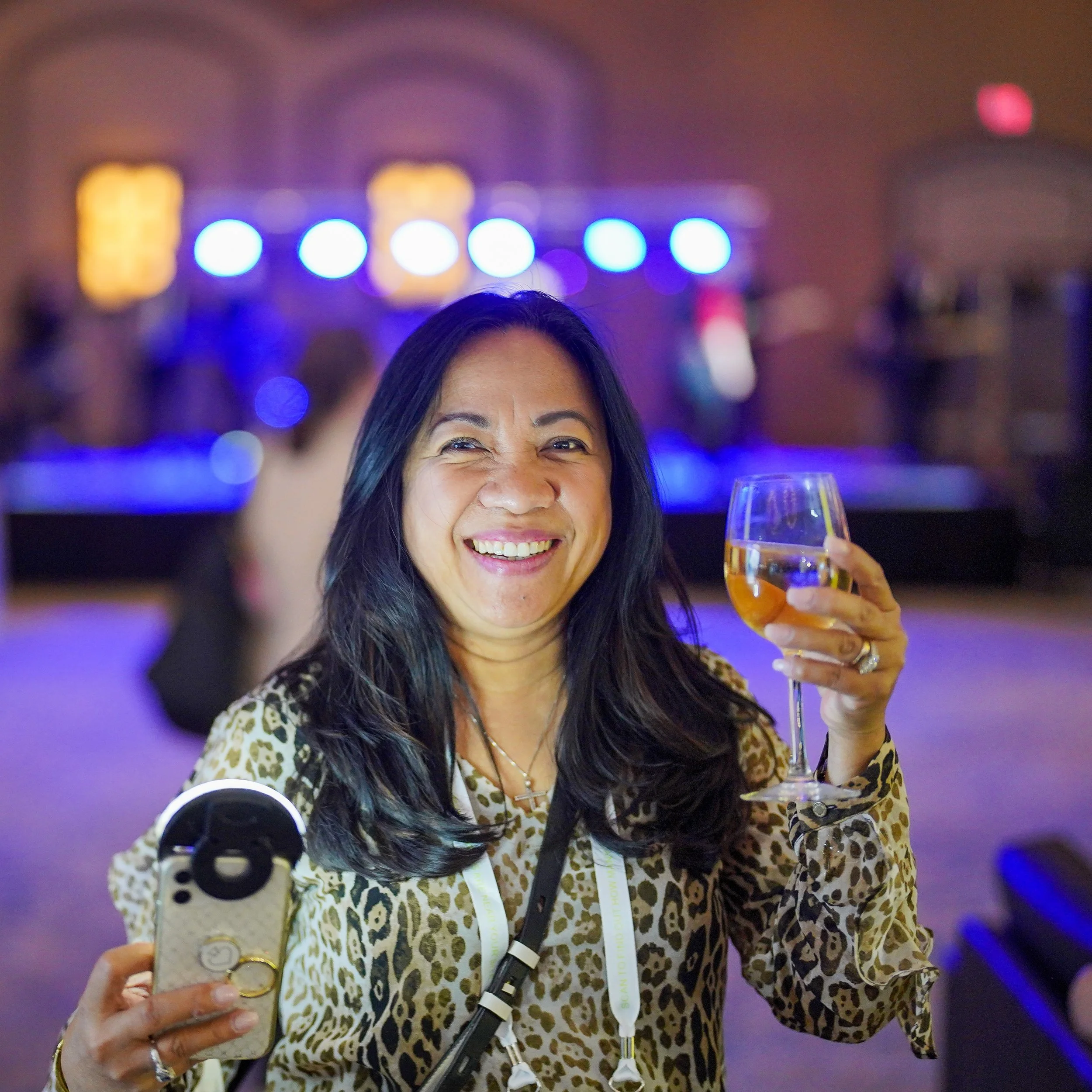 A woman with long dark hair smiling and holding a glass of wine at an indoor event with a stage and blue lights in the background.