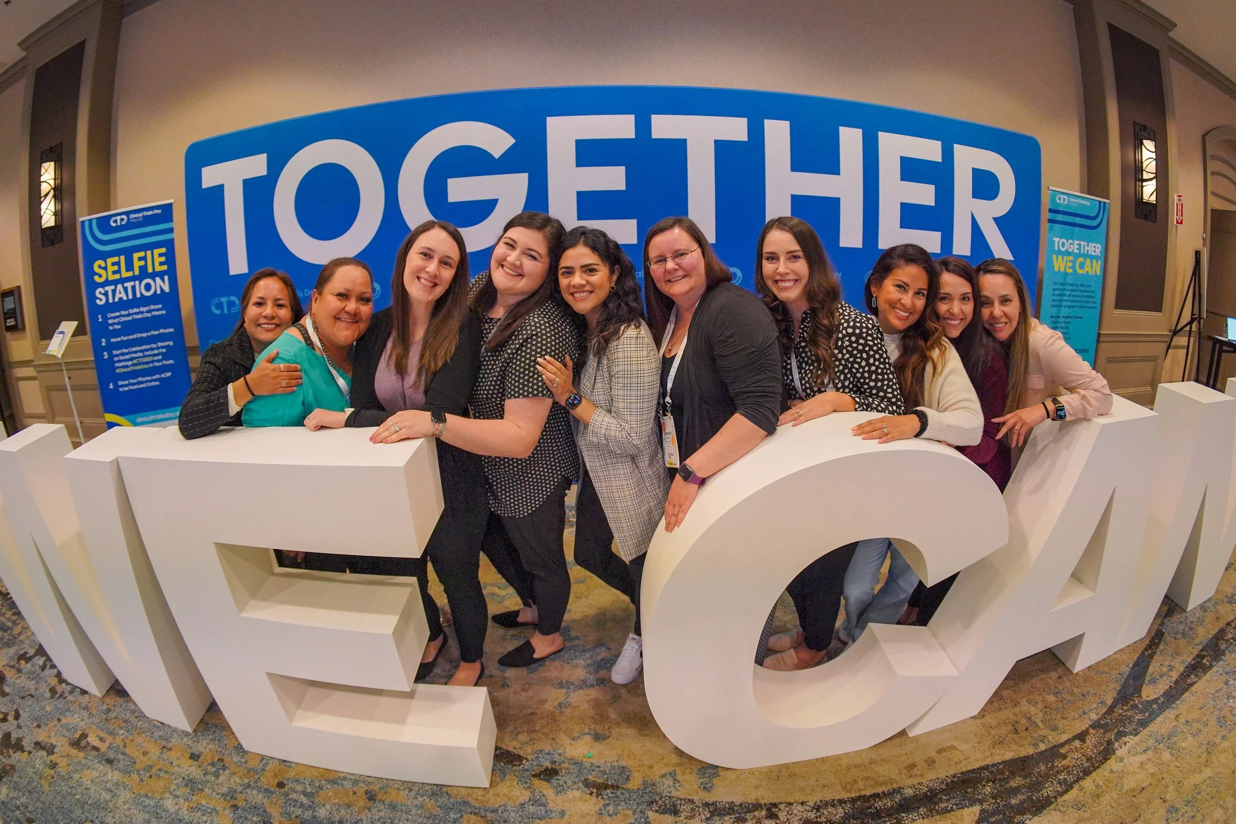 Group of women smiling and posing behind large white letters spelling 'EDUCATION' at a conference with a blue and white 'Together We Can' backdrop and a selfie station sign.