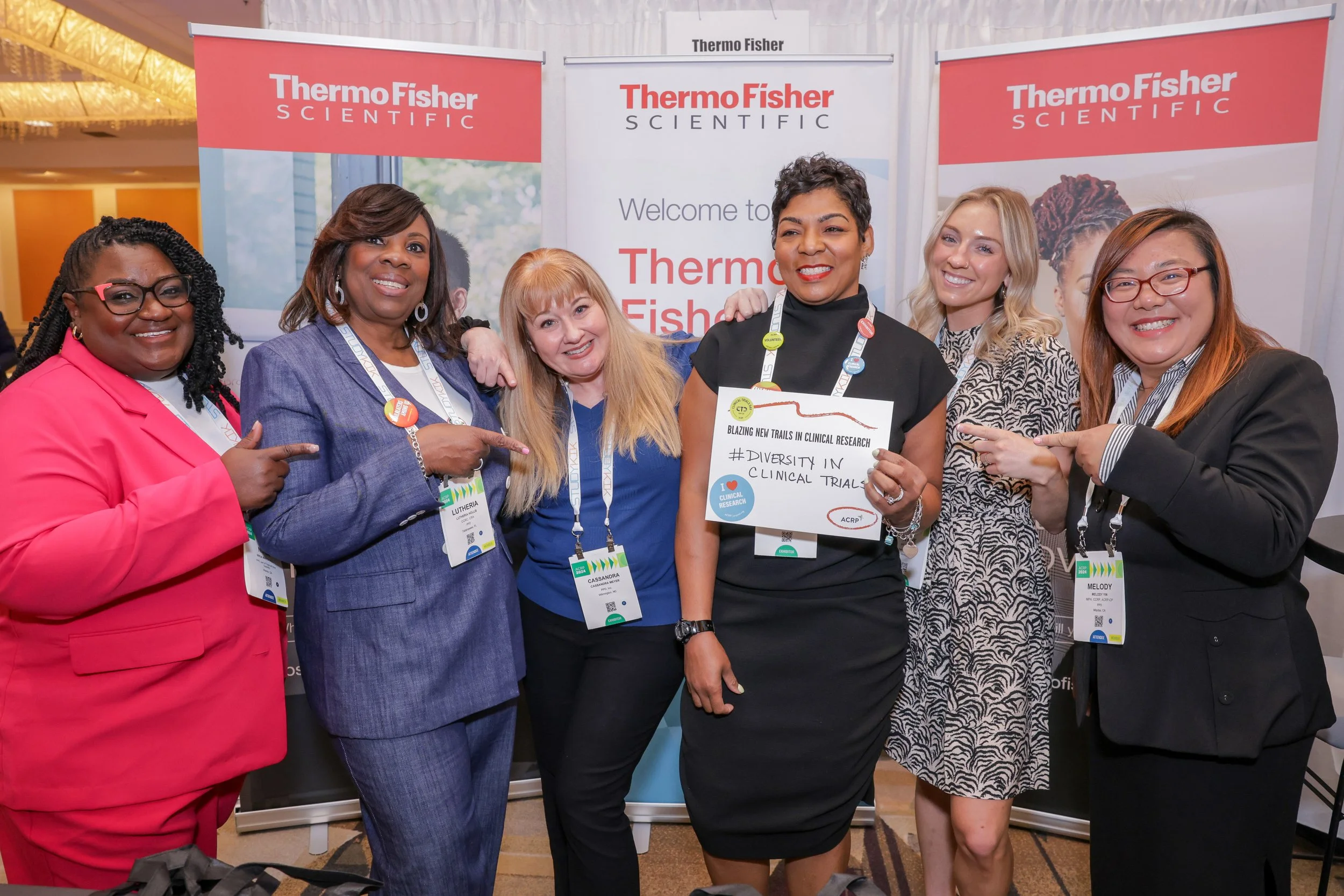 Group of six women at a conference, standing in front of Thermo Fisher Scientific banners, smiling, some pointing at a woman holding a sign that says 'Diversity in Clinical Trials'.