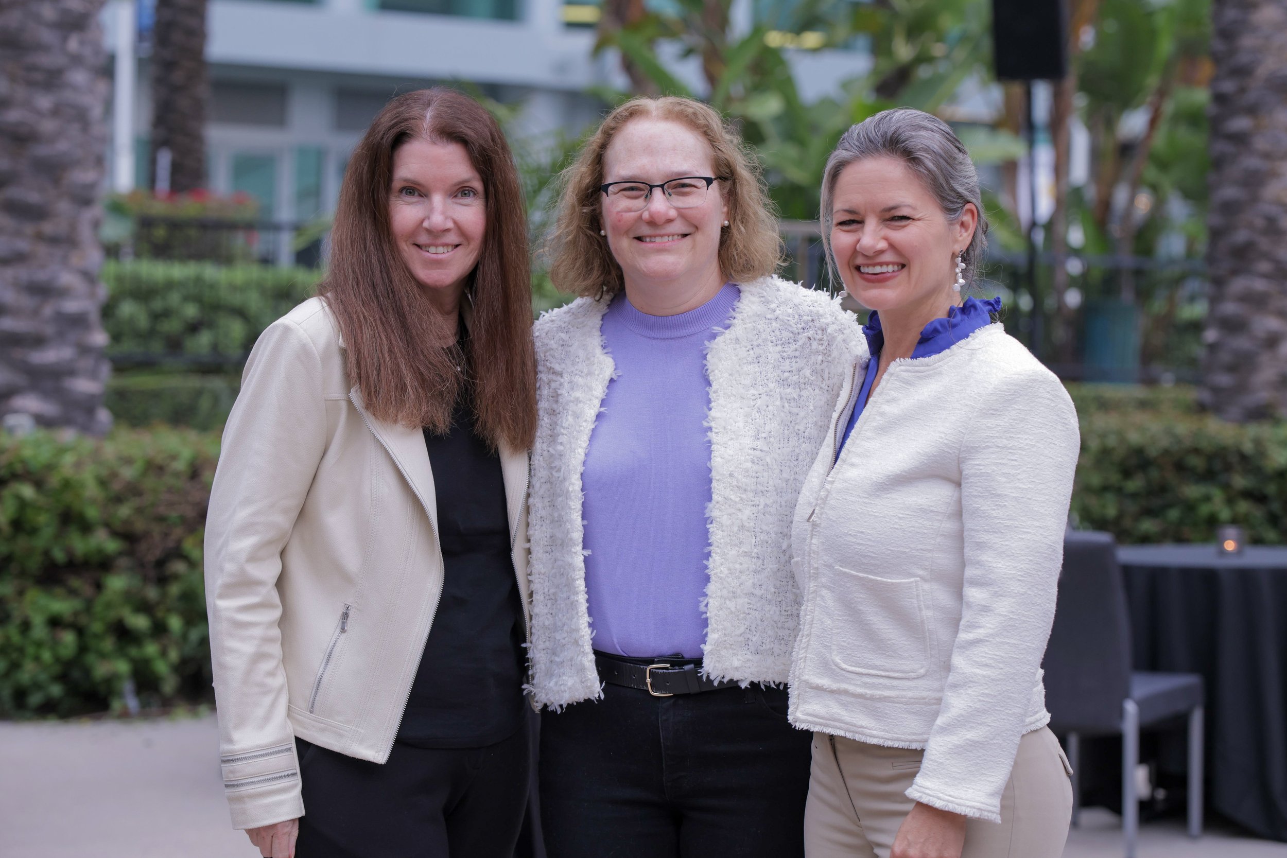 Three women standing outdoors, smiling for a photo. They are dressed in light-colored jackets and are surrounded by green plants and trees.