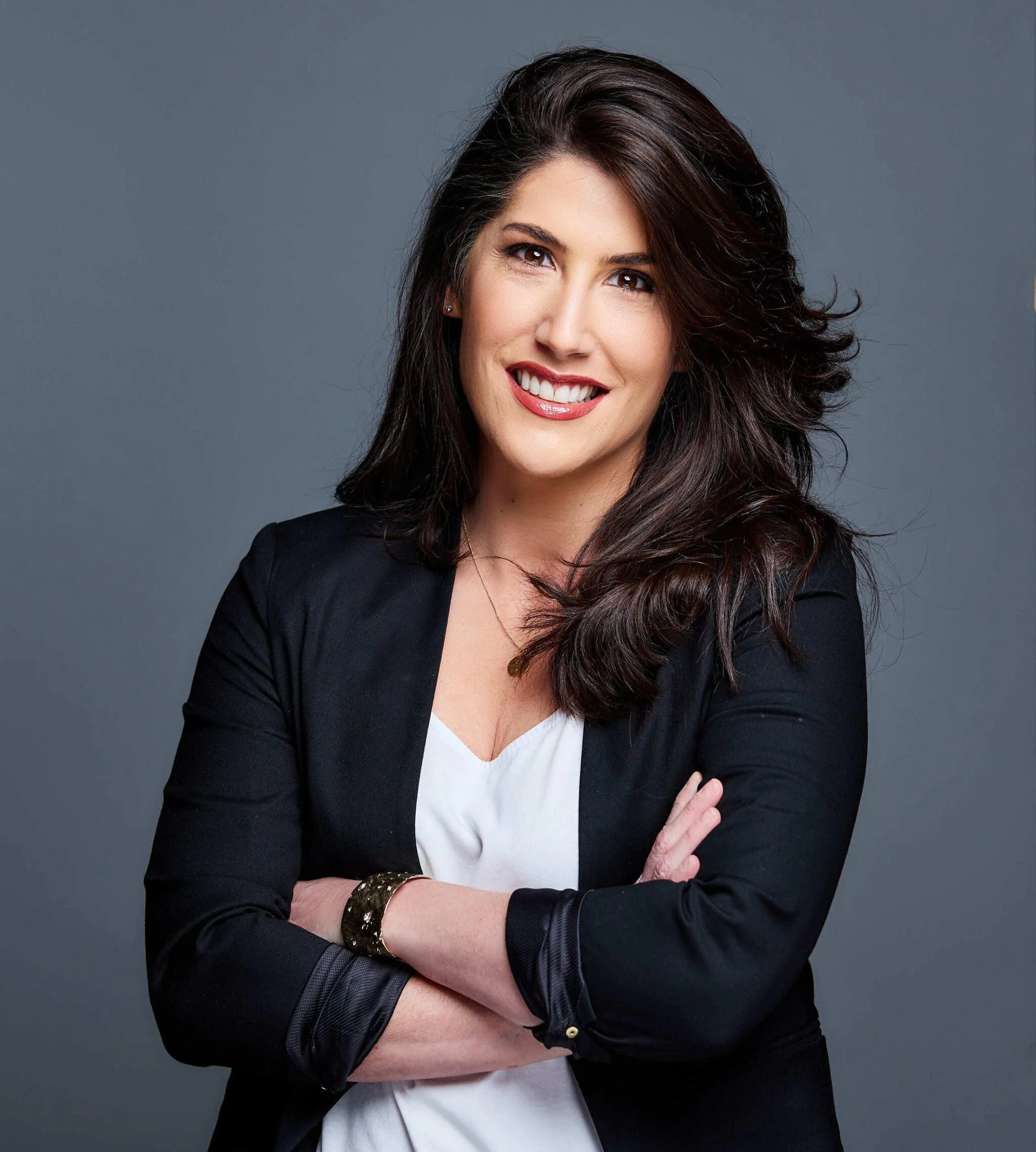Close-up portrait of a woman with long dark brown hair, smiling, wearing a black top, against a plain light gray background.
