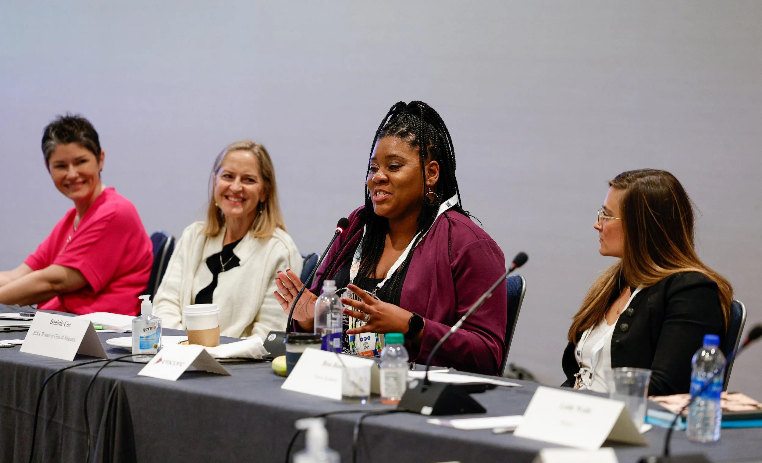 Four women sitting at a panel discussion table, with one woman speaking into a microphone, in a conference room setting.
