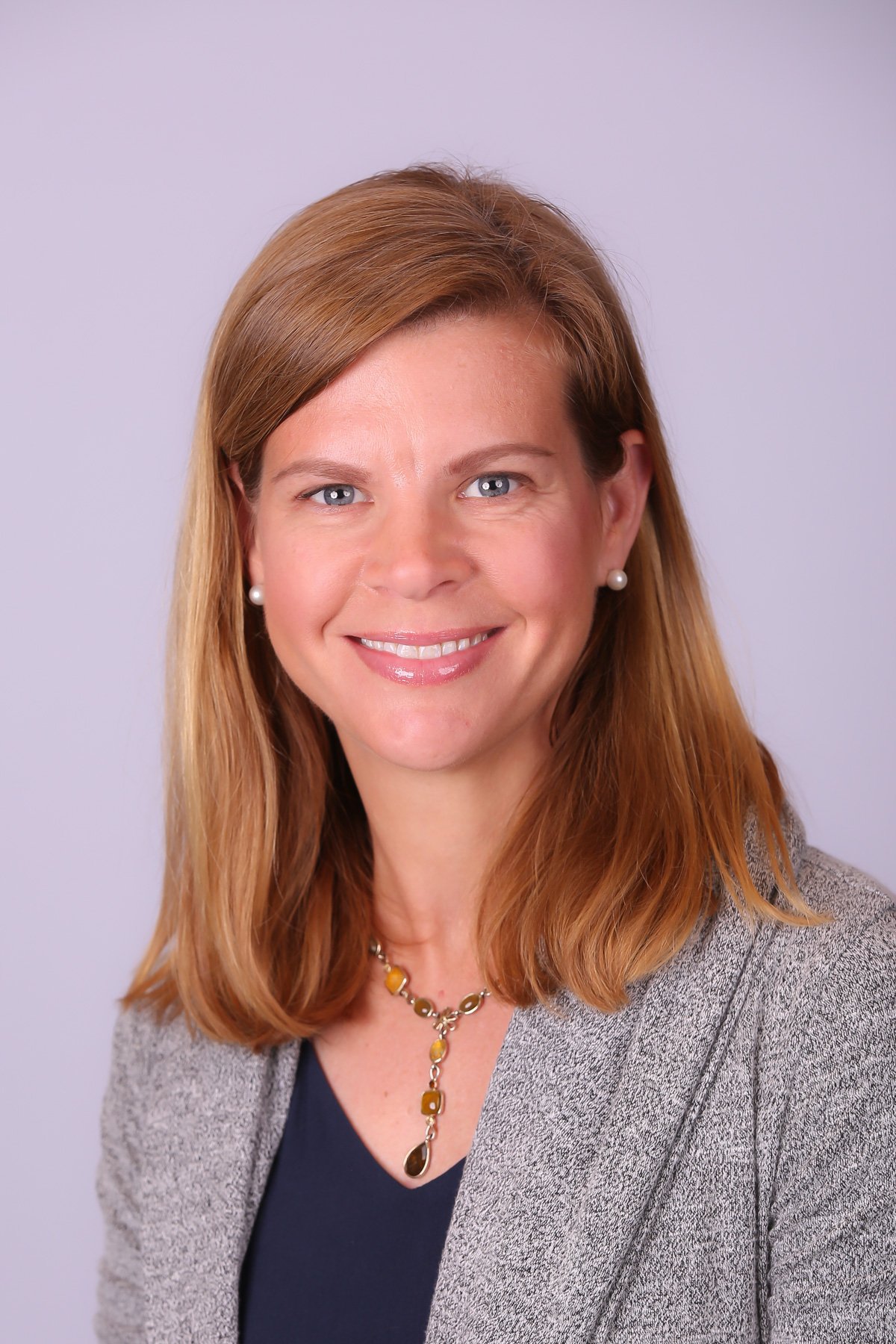 A portrait of a woman with shoulder-length red hair, wearing a gray blazer, navy top, pearl earrings, and a necklace with yellow and brown stones, smiling against a plain light background.