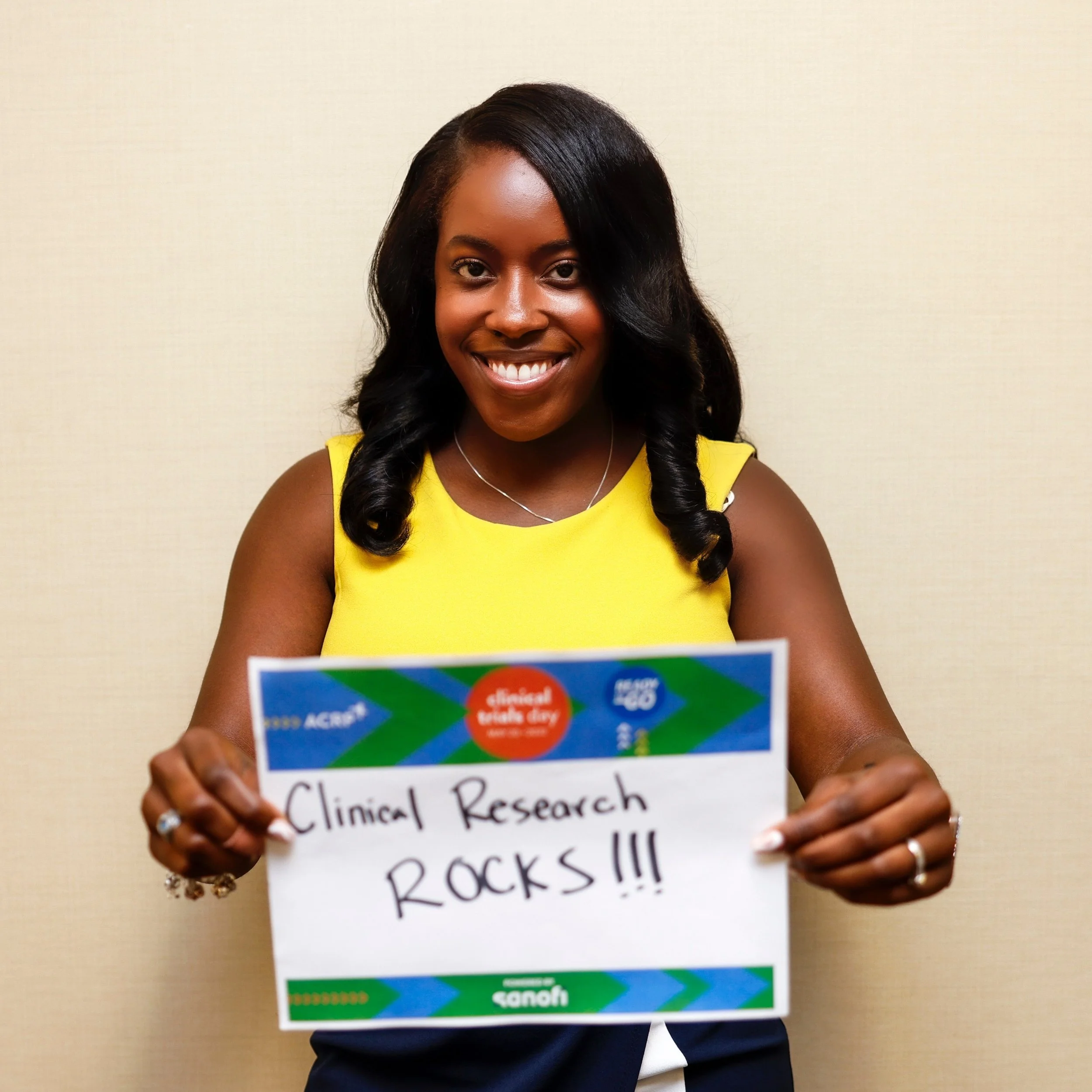 Smiling woman in a yellow top holding a sign that says "Clinical Research ROCKS!!!".