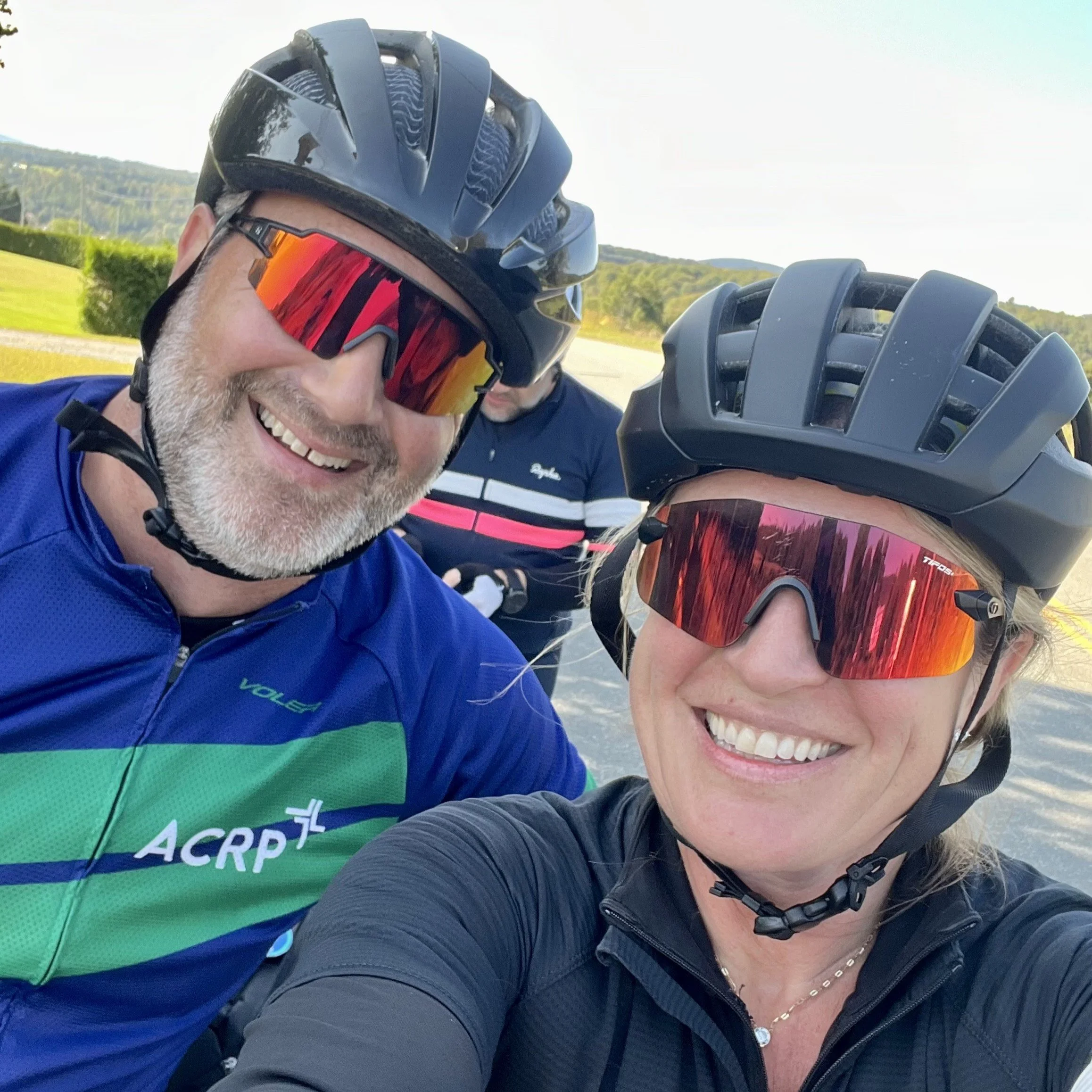 Two smiling adults wearing cycling helmets and sunglasses taking a selfie outdoors on a sunny day with a man in the background.