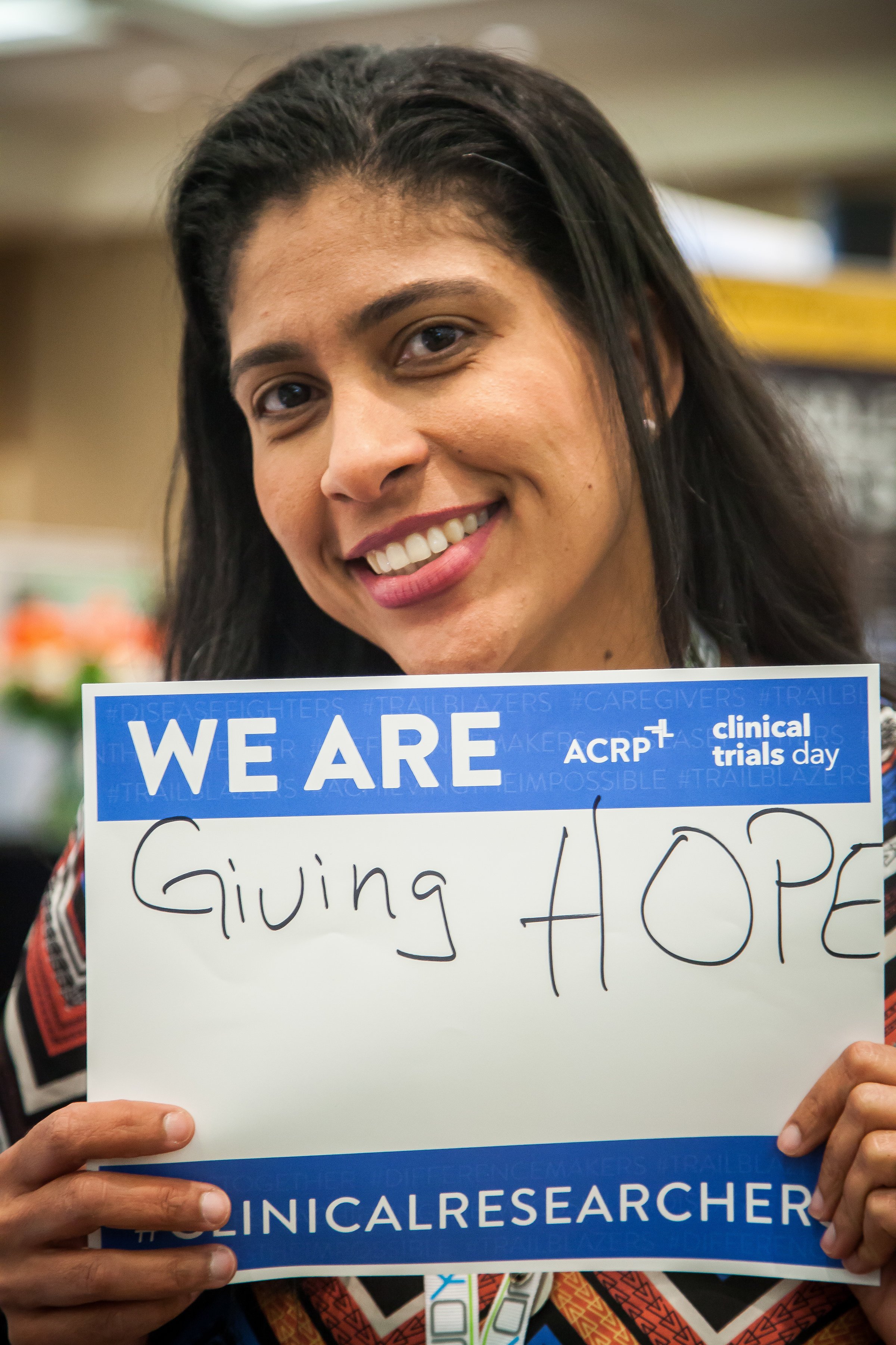 A smiling woman holding a sign that reads "WE ARE Giving HOPE" at a clinical trials event.