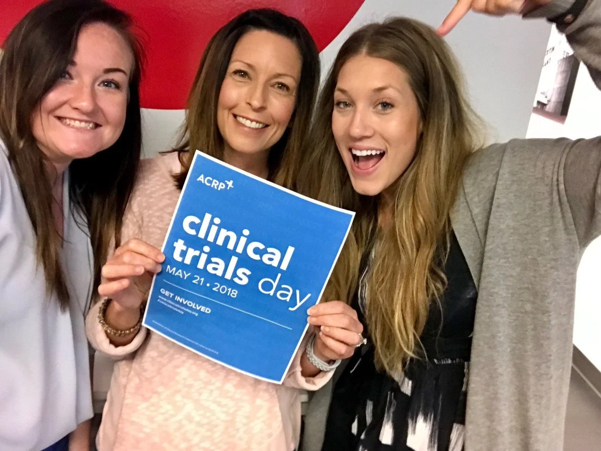 Three women smiling at a medical conference, holding a blue sign that reads 'clinical trials day May 21, 2018.' The woman in the middle has light brown hair and is holding the sign, while the woman on the right has long, wavy blonde hair, and the wom
