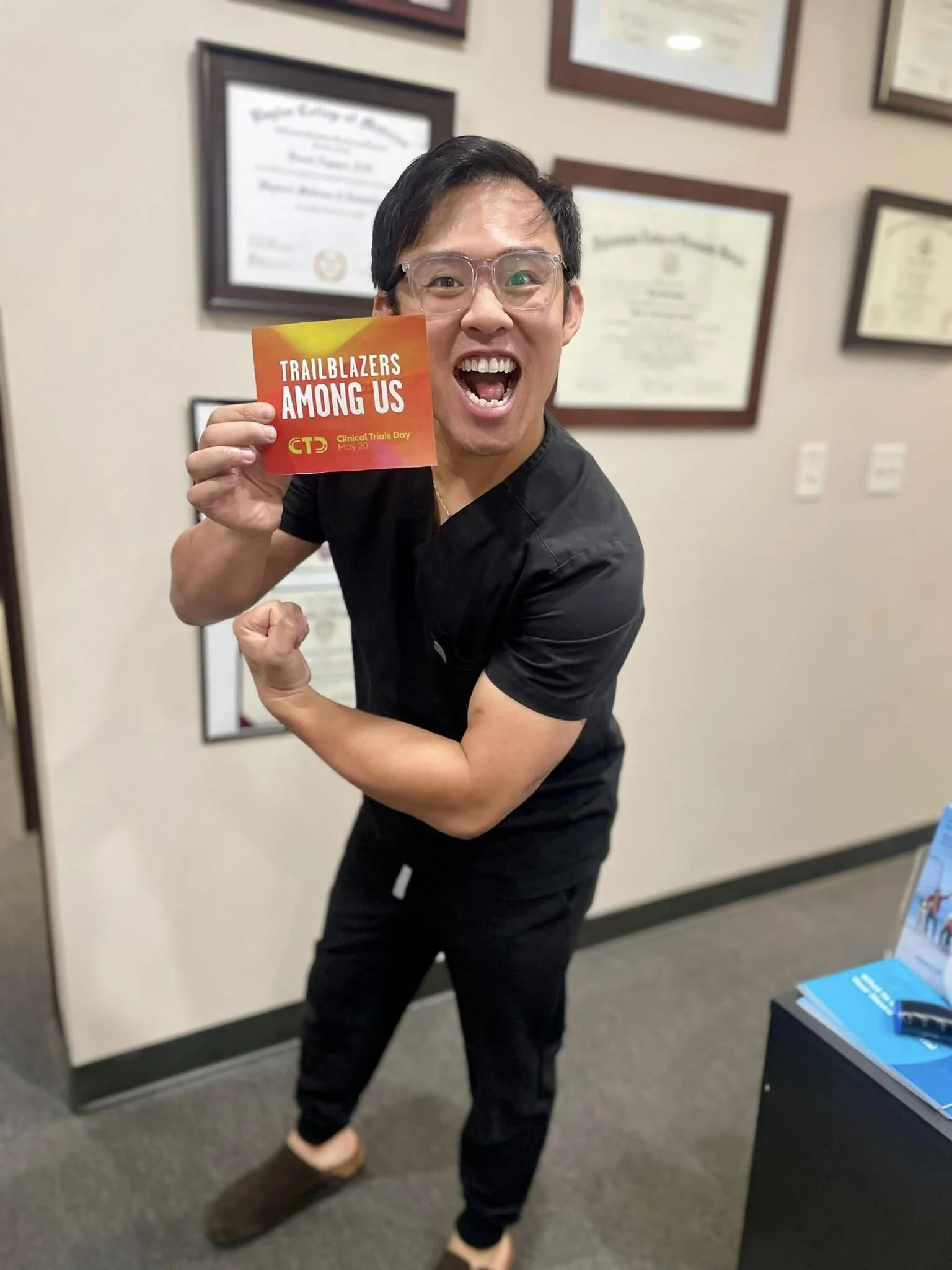 A man in black medical scrubs and glasses is smiling widely, holding a red and yellow card that reads 'Trailblazers Among Us. Clinical Trials Day May 20'. He is flexing his right arm, showing muscle, and standing inside an office with framed certific