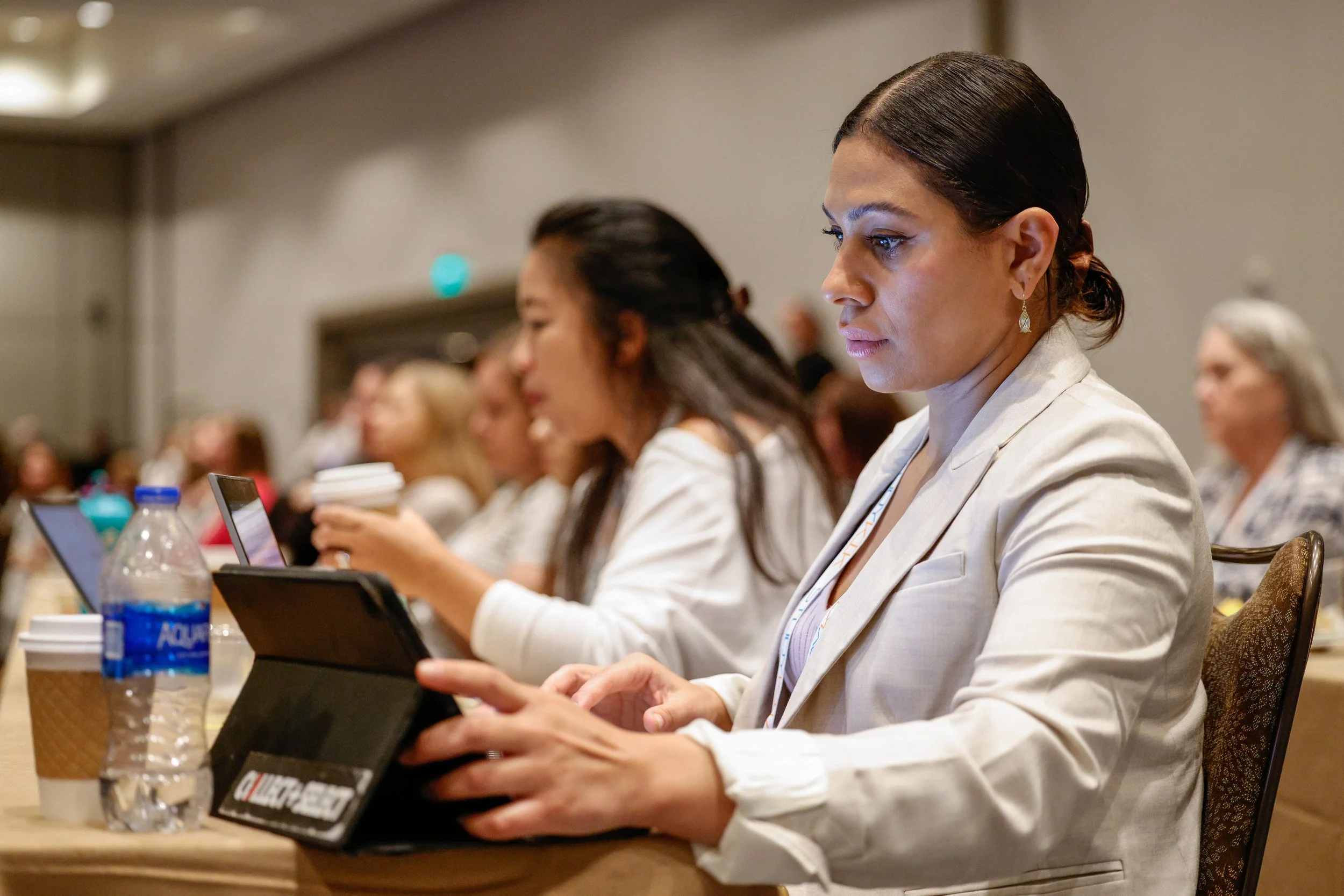 A woman in a white blazer sitting at a conference table, looking attentively at her tablet device, with other women in professional attire seated beside her, some using laptops or holding coffee cups.