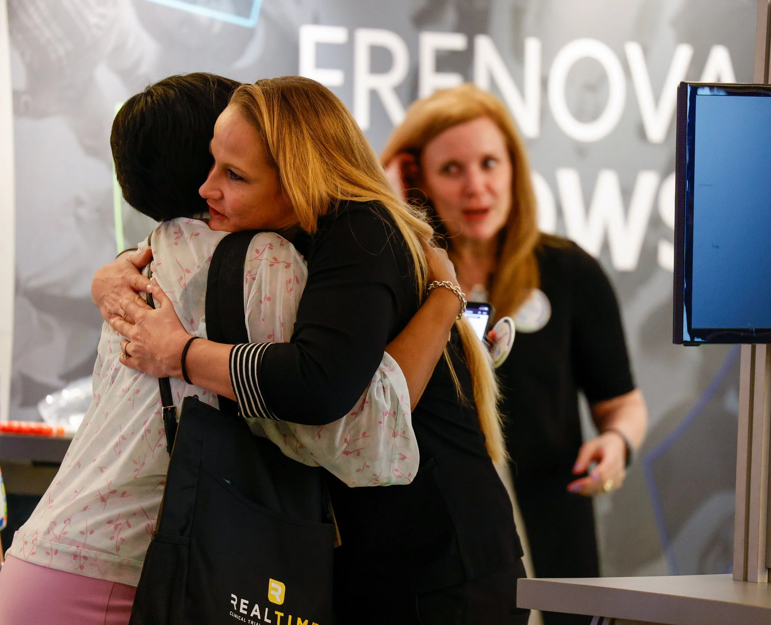 Two women hugging at an event, one woman with blonde hair and black clothing, another woman with dark hair and patterned clothing. A third woman with long blonde hair is visible in the background.
