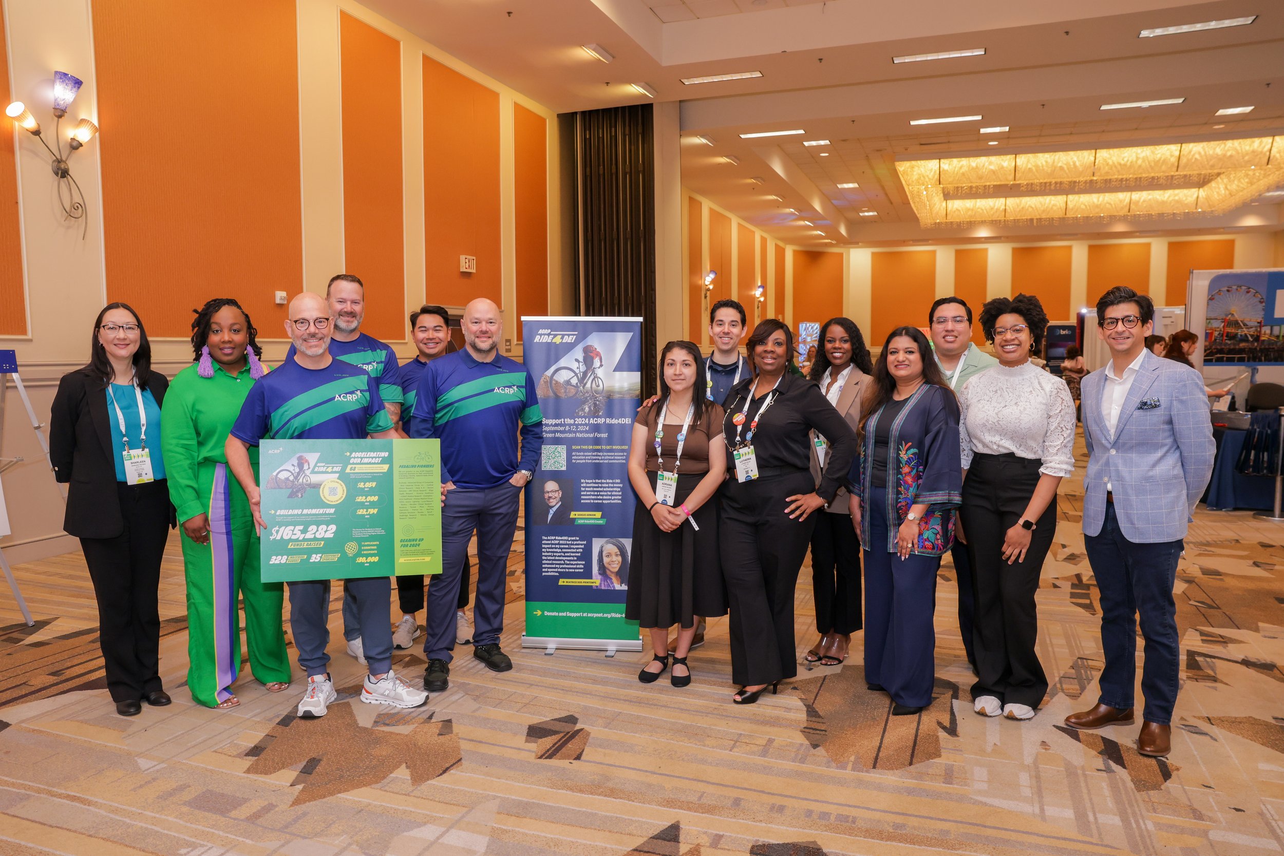 A group of diverse people standing together in a banquet hall, some holding a large poster board and a vertical banner promoting a cycling event or fundraiser, with colorful wall panels and artworks in the background.