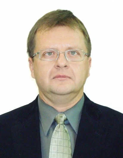 A portrait of a middle-aged man with glasses, wearing a dark suit, gray shirt, and patterned tie, against a plain white background.