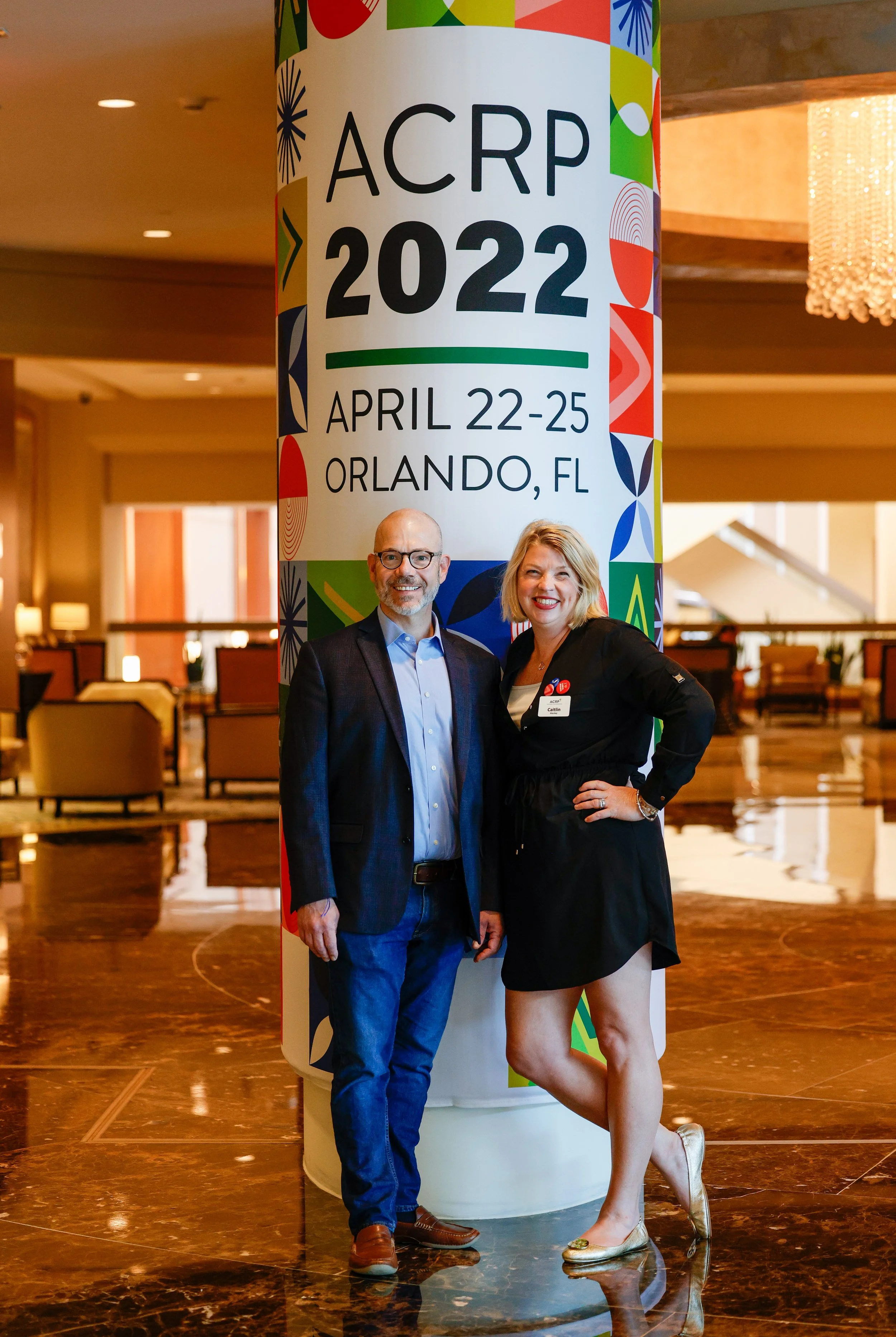 Two people standing in front of a large colorful banner at the ACRP 2022 conference, which took place from April 22-25 in Orlando, Florida. The man is wearing a suit and glasses, and the woman is wearing a black dress with a name badge.