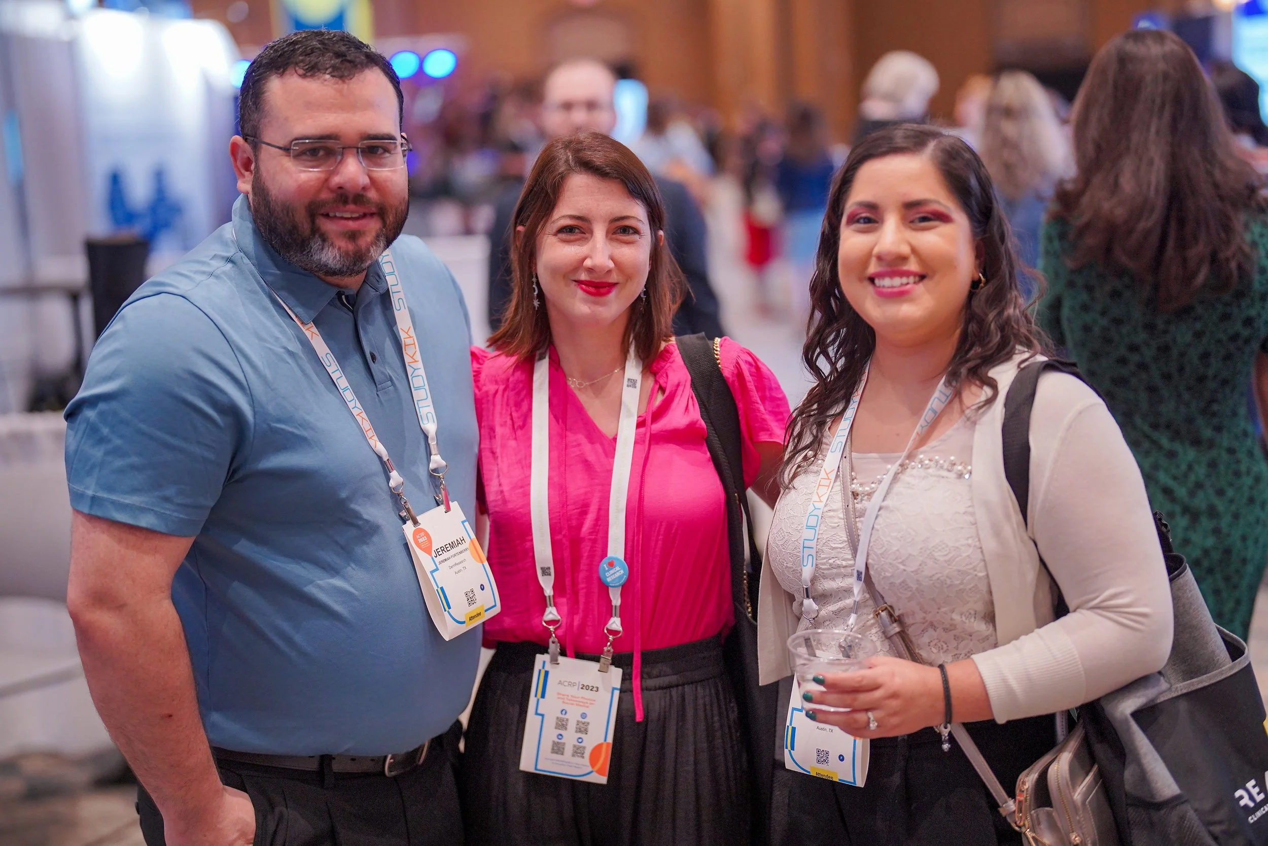 Three people smiling and standing together at a conference, wearing lanyards with name tags, with a busy event hall in the background.