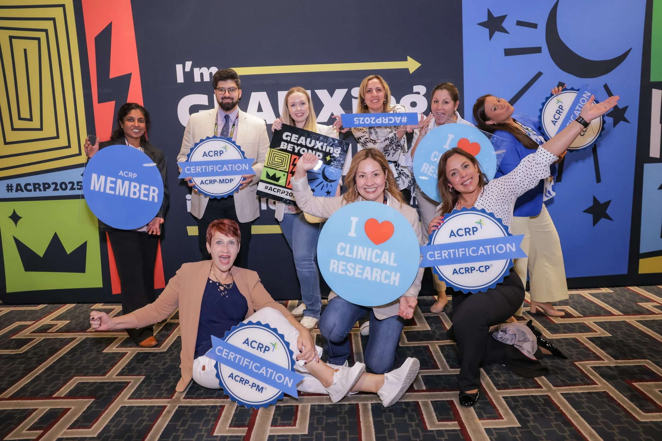 Group of nine diverse people smiling and holding signs at a professional conference or event. Signs include phrases like 'ACRP MEMBER,' 'CERTIFICATION,' 'I ❤️ CLINICAL RESEARCH,' and other certification-related messages. The background features color