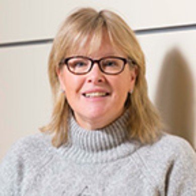 A woman with blonde hair, glasses, and a gray turtleneck sweater smiling indoors.