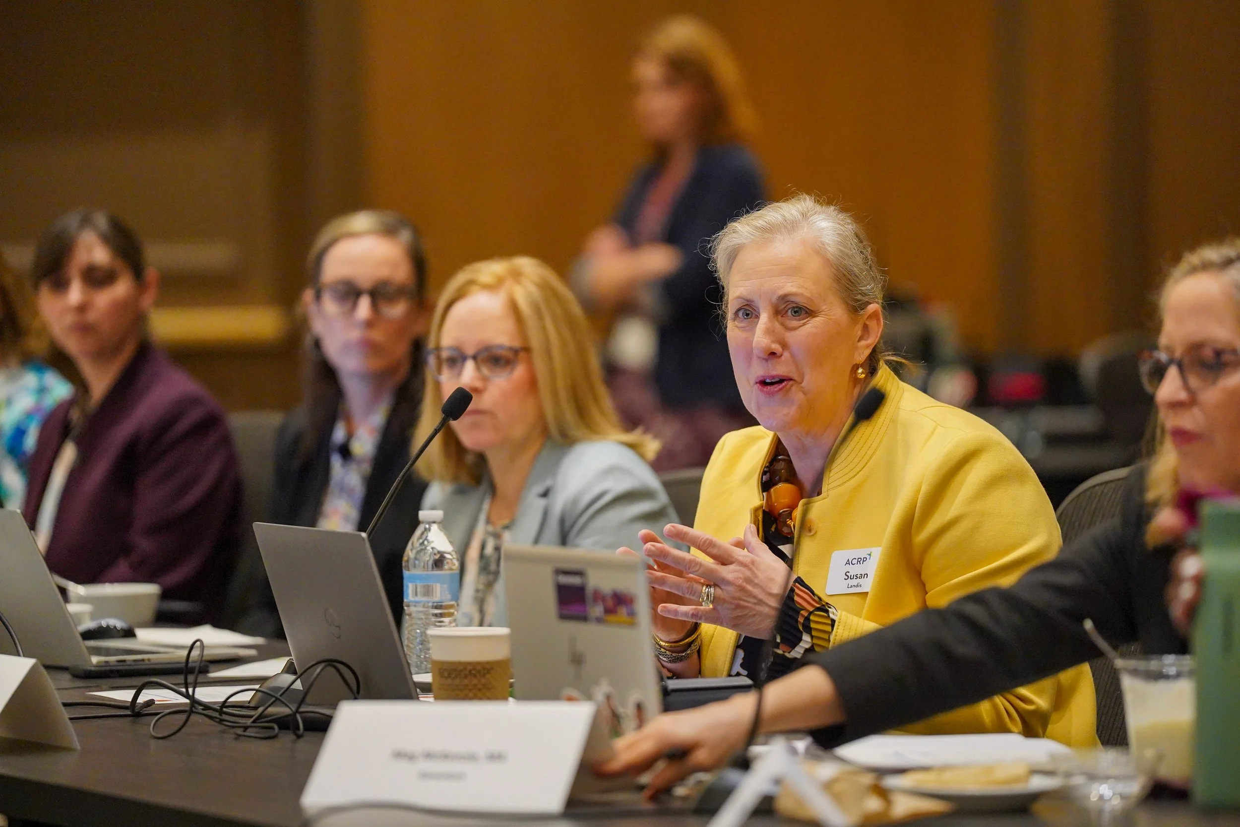 A woman in a yellow blazer speaking at a conference table with others listening, in a formal meeting room.