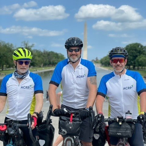 Three cyclists in matching blue and white jerseys, wearing helmets and sunglasses, standing with their bikes outdoors on a sunny day with a canal and trees in the background.