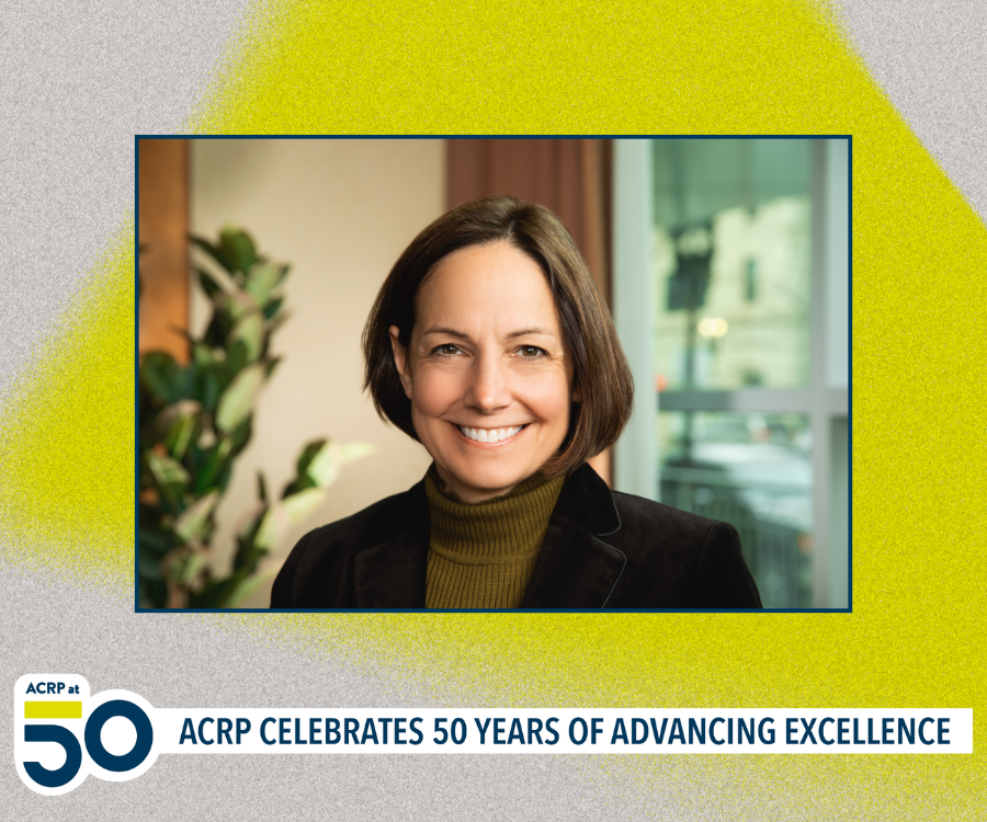 A woman with short brown hair smiling in a video frame set in an indoor setting with a plant and window in the background.
