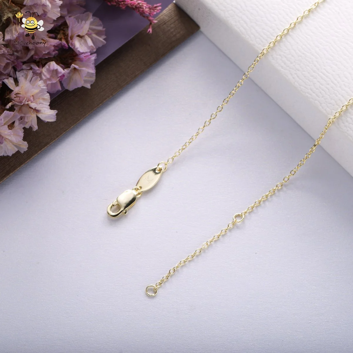 Close-up of a gold necklace with a clasp, resting on a white surface with purple flowers on a wooden tray in the background.